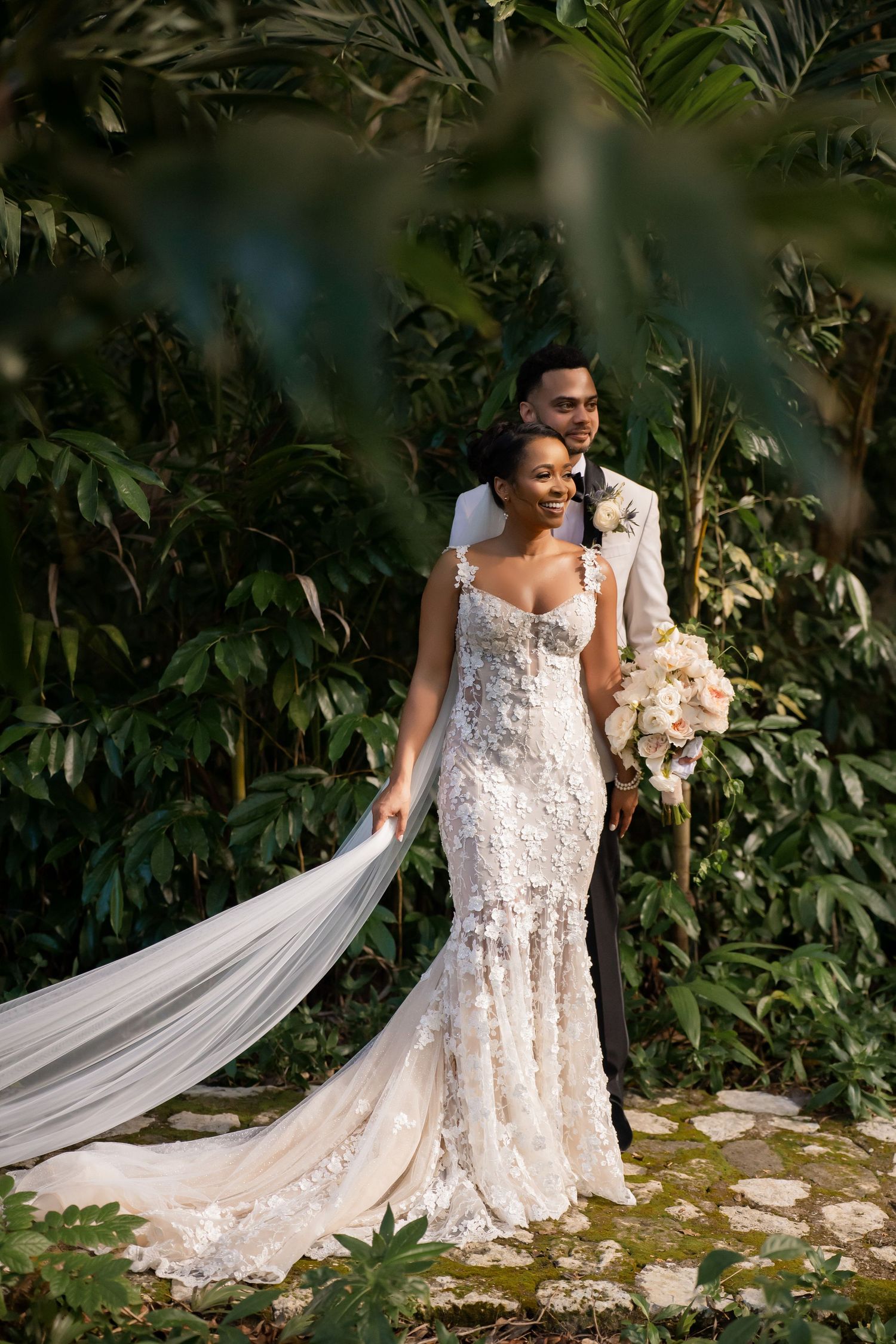 A romantic wedding couple poses together outdoors among lush tropical greenery with a flowing white lace gown and long veil.