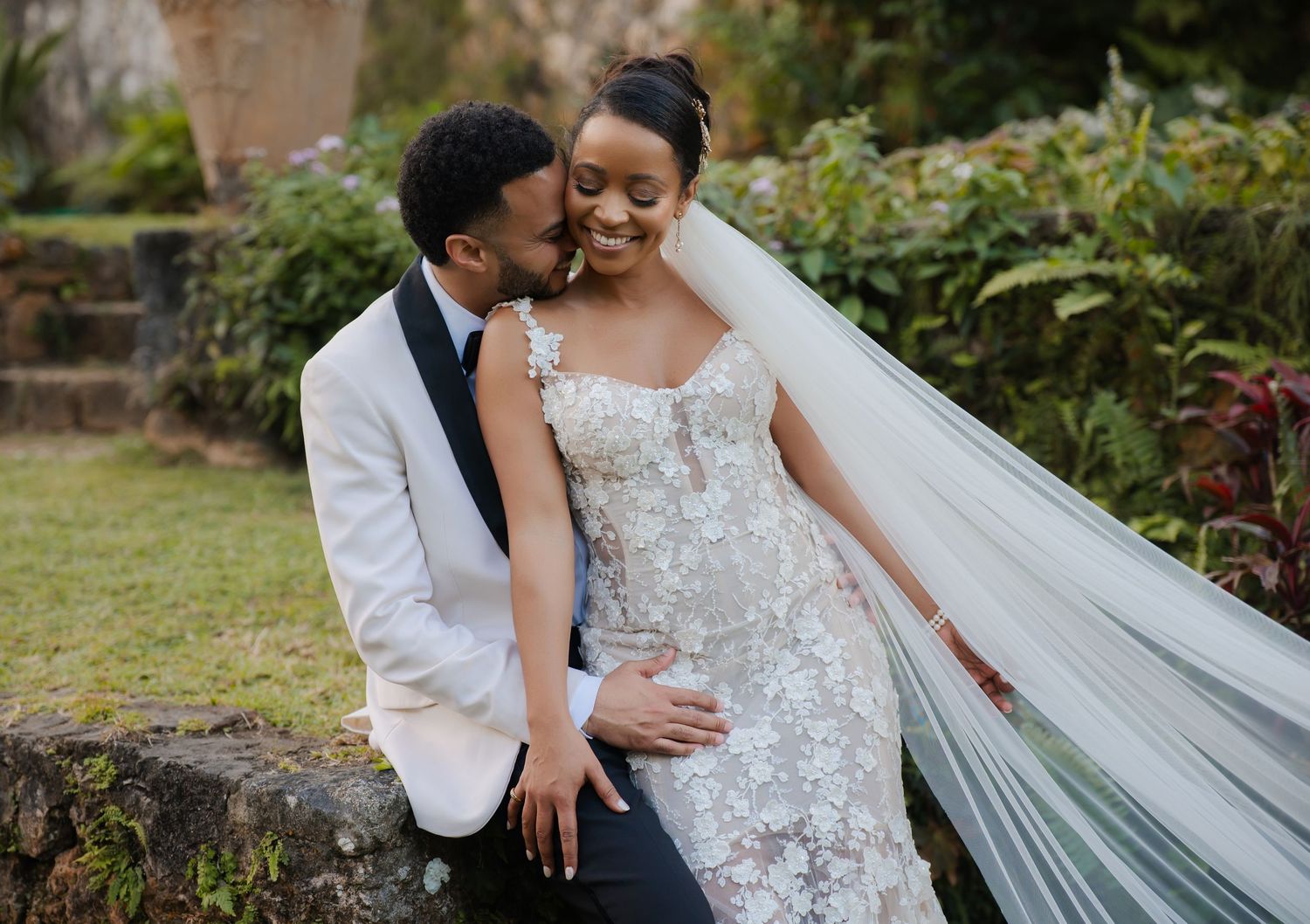 Couple shares intimate moment outdoors on their wedding day with bride wearing lace gown and flowing veil.