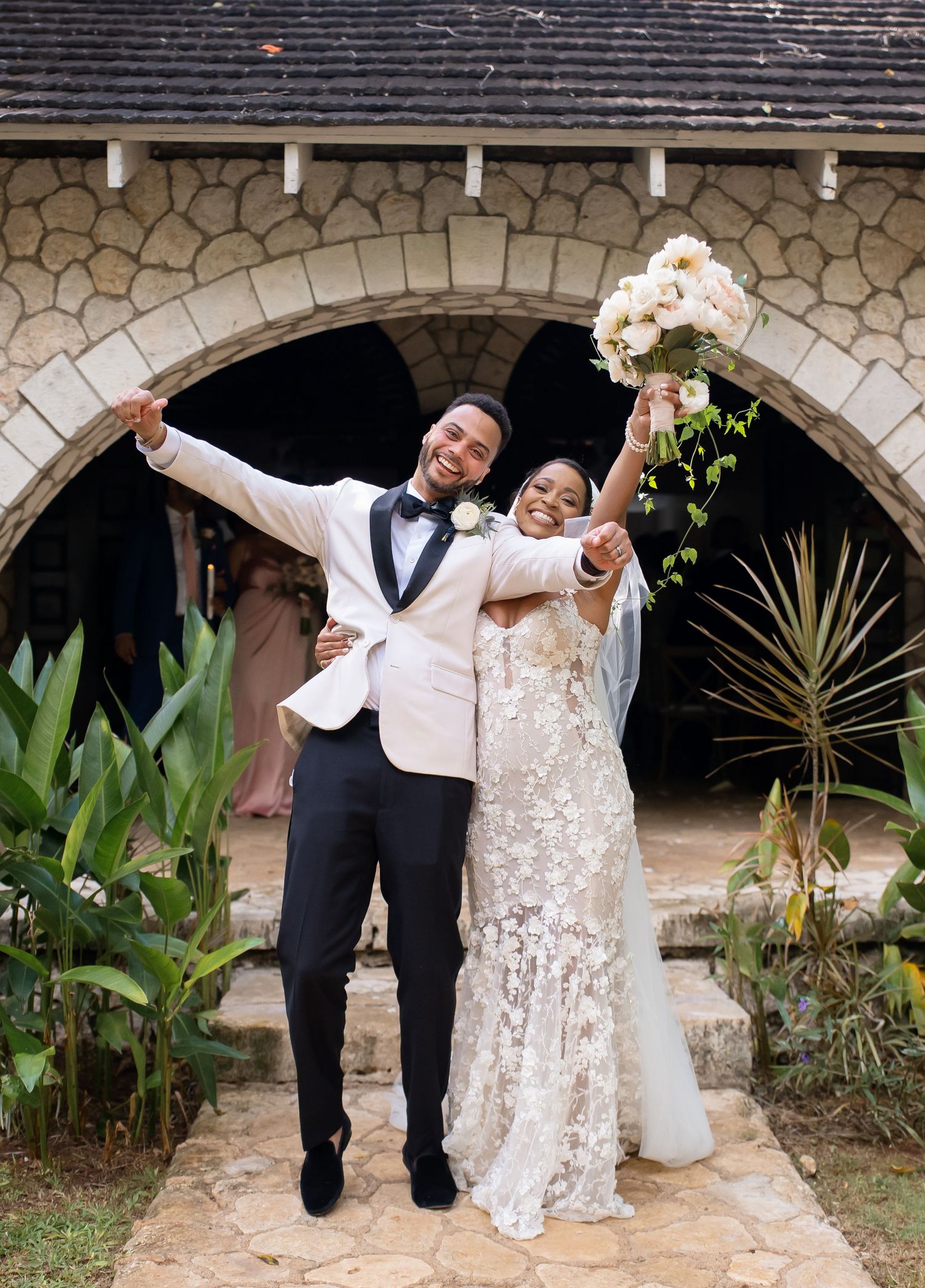 Joyful couple celebrates their wedding day in front of a stone archway surrounded by tropical plants.