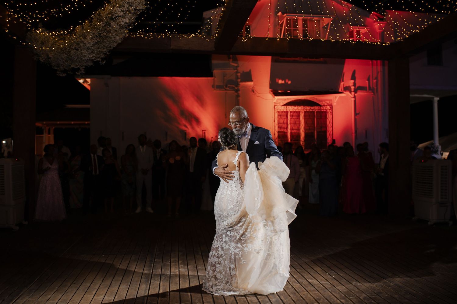 A couple in long white dresses dances together on a dark dance floor illuminated by red ambient lighting.