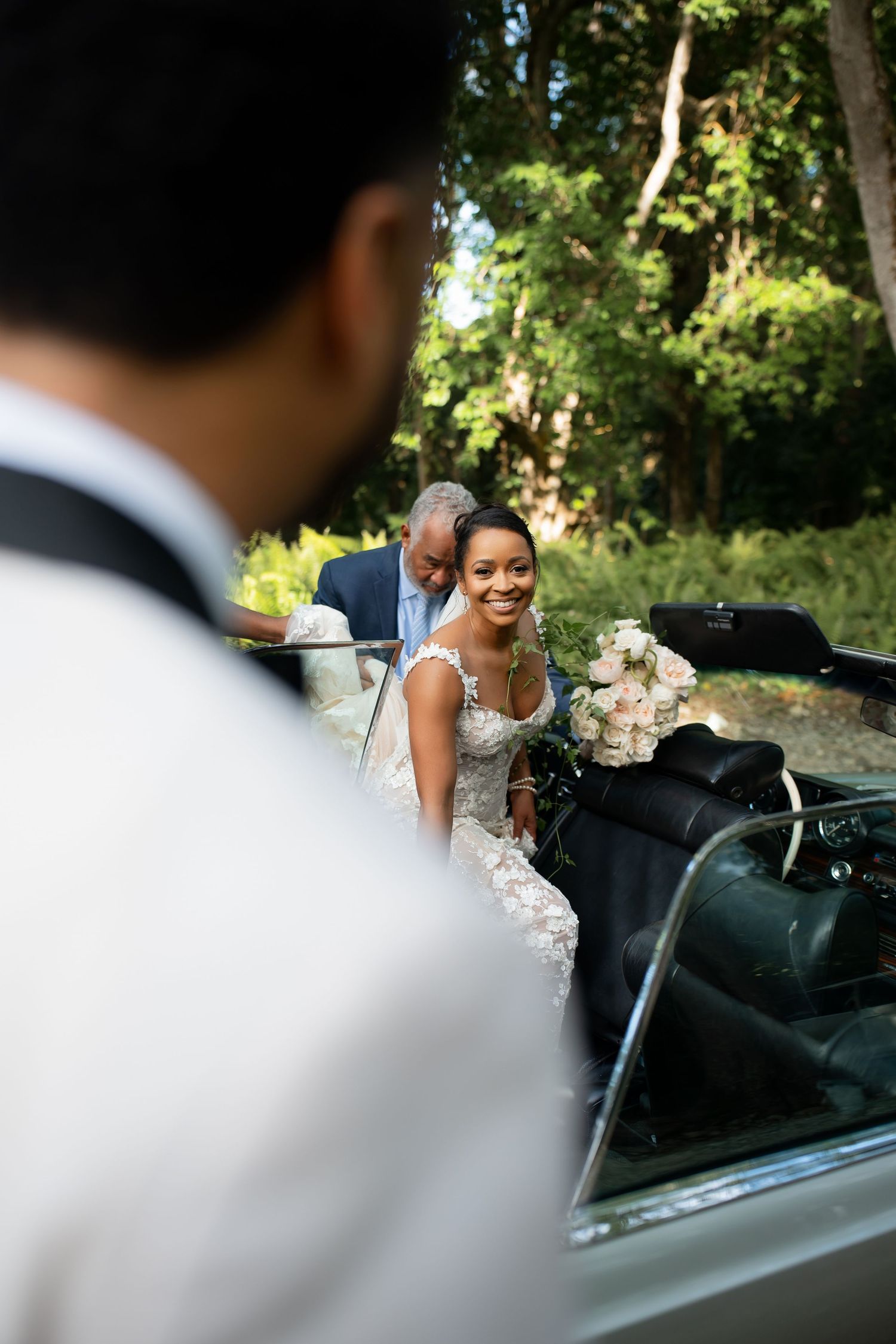 A couple embraces and smiles while leaning over a vintage black convertible car in a sunny outdoor setting.