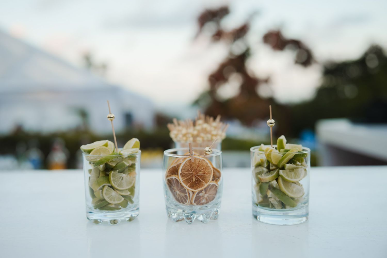 Three decorative cocktail glasses with fresh lime garnishes and herbs arranged on a white surface at outdoor event.