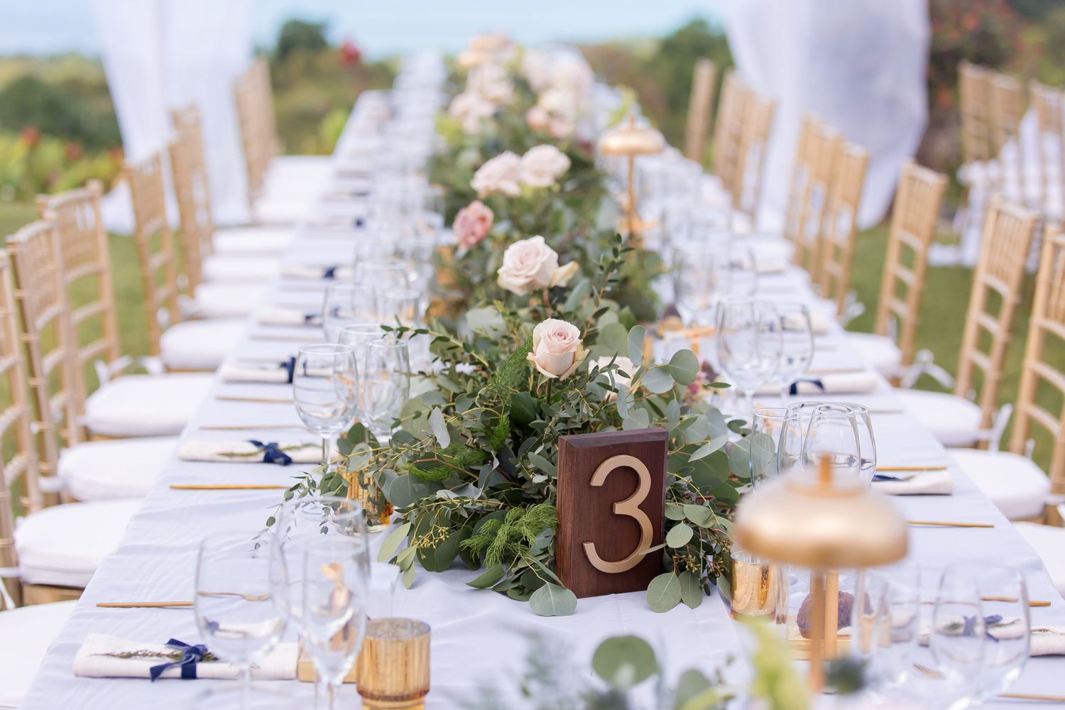 An elegant wedding reception table setting with white linens, greenery garlands, and golden table number accents.