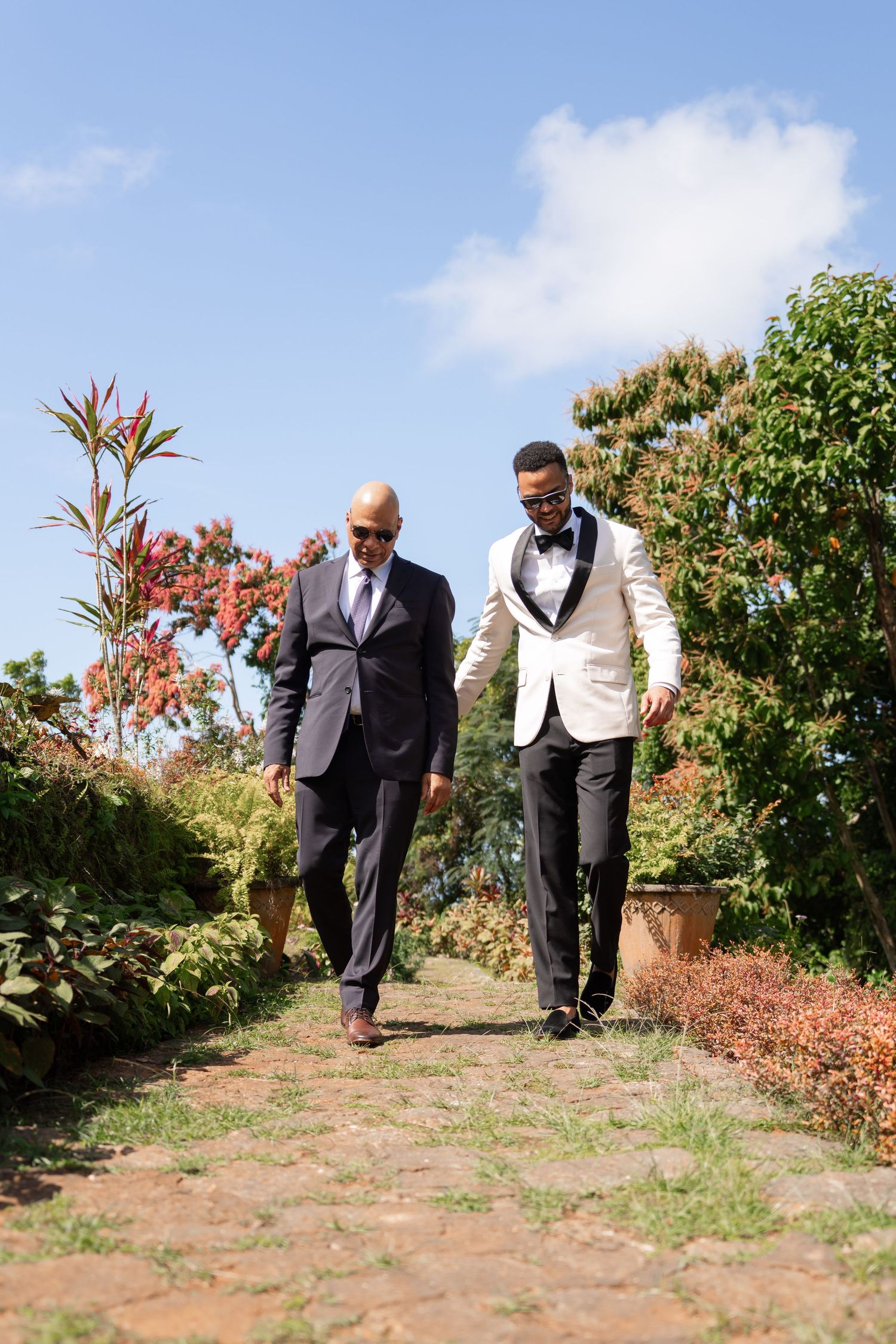 Two people in formal attire examine plants in an outdoor garden with blue sky and shrubs in the background.