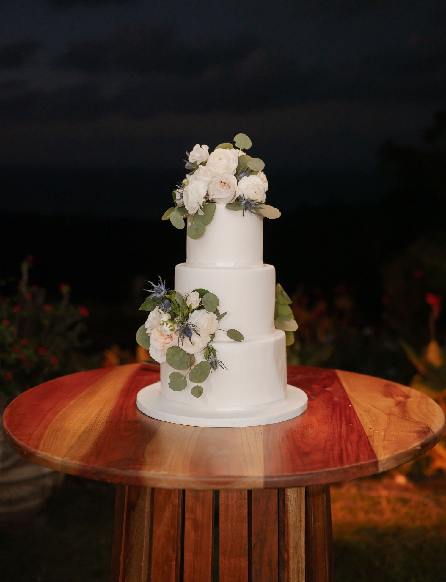 Elegant three-tier white wedding cake decorated with blush roses and eucalyptus leaves sits on wooden round table.