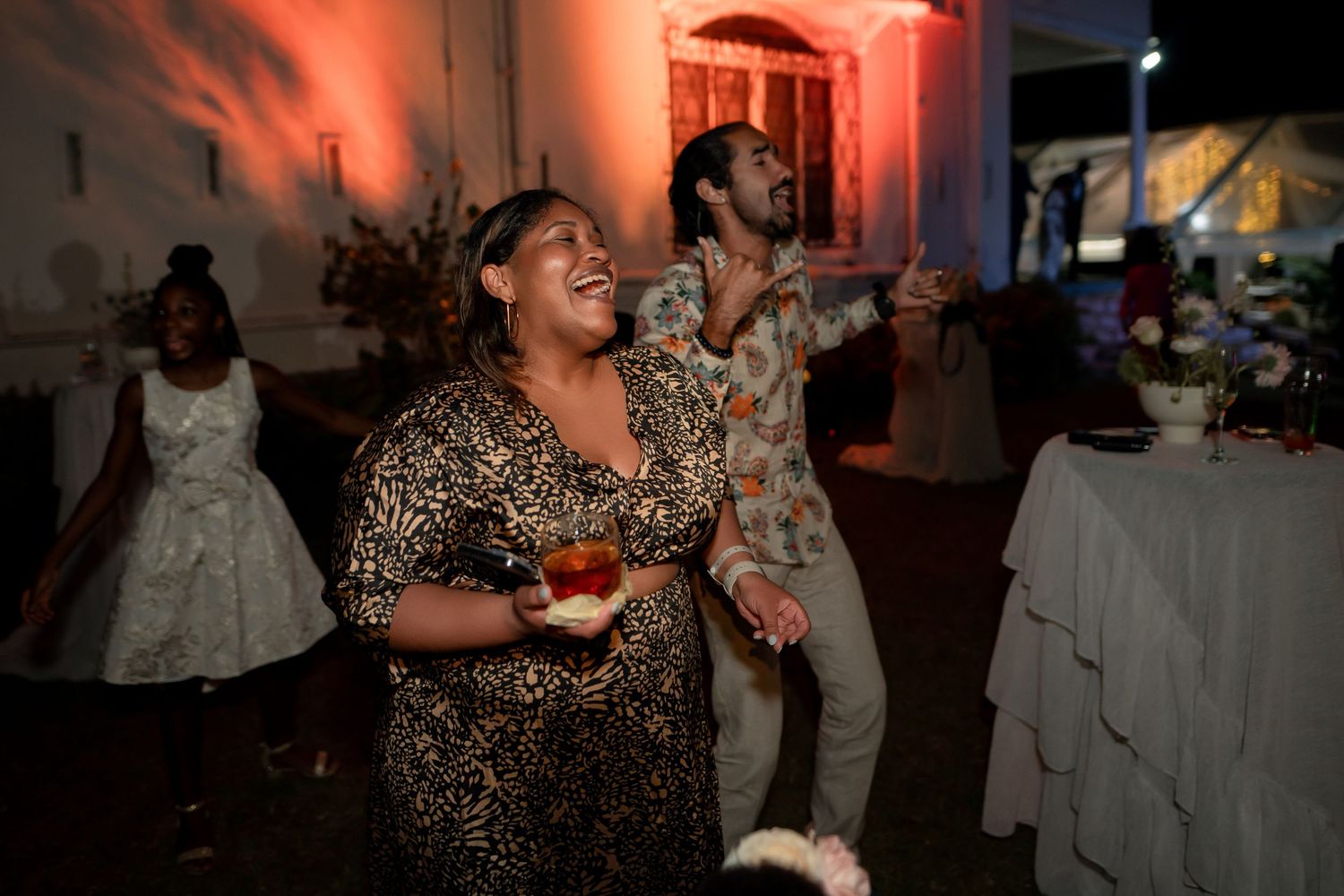 People dancing and enjoying themselves at an evening party with red ambient lighting and white tablecloths.