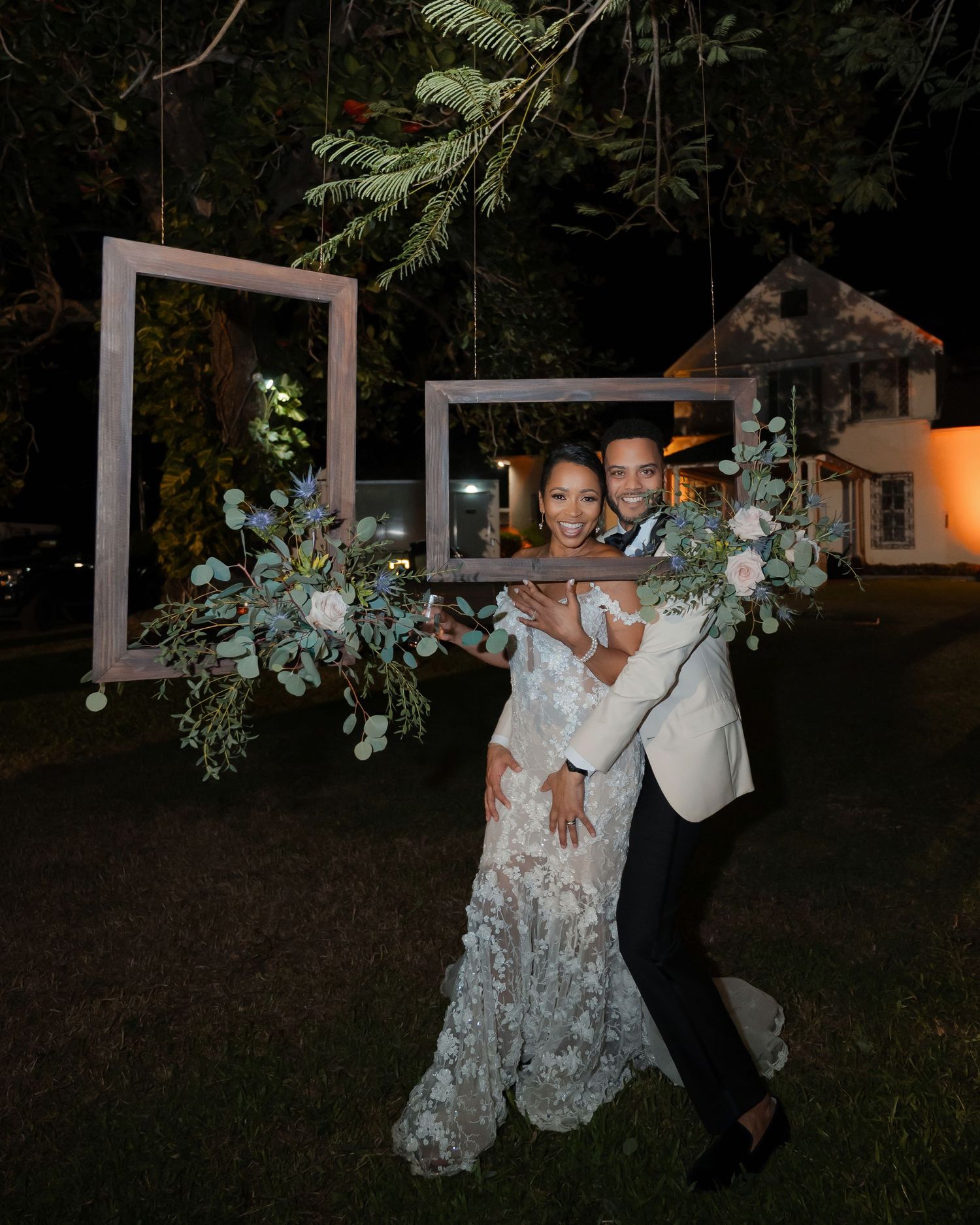 A couple poses in an outdoor wedding setup with a suspended white frame decorated with greenery and florals at night.
