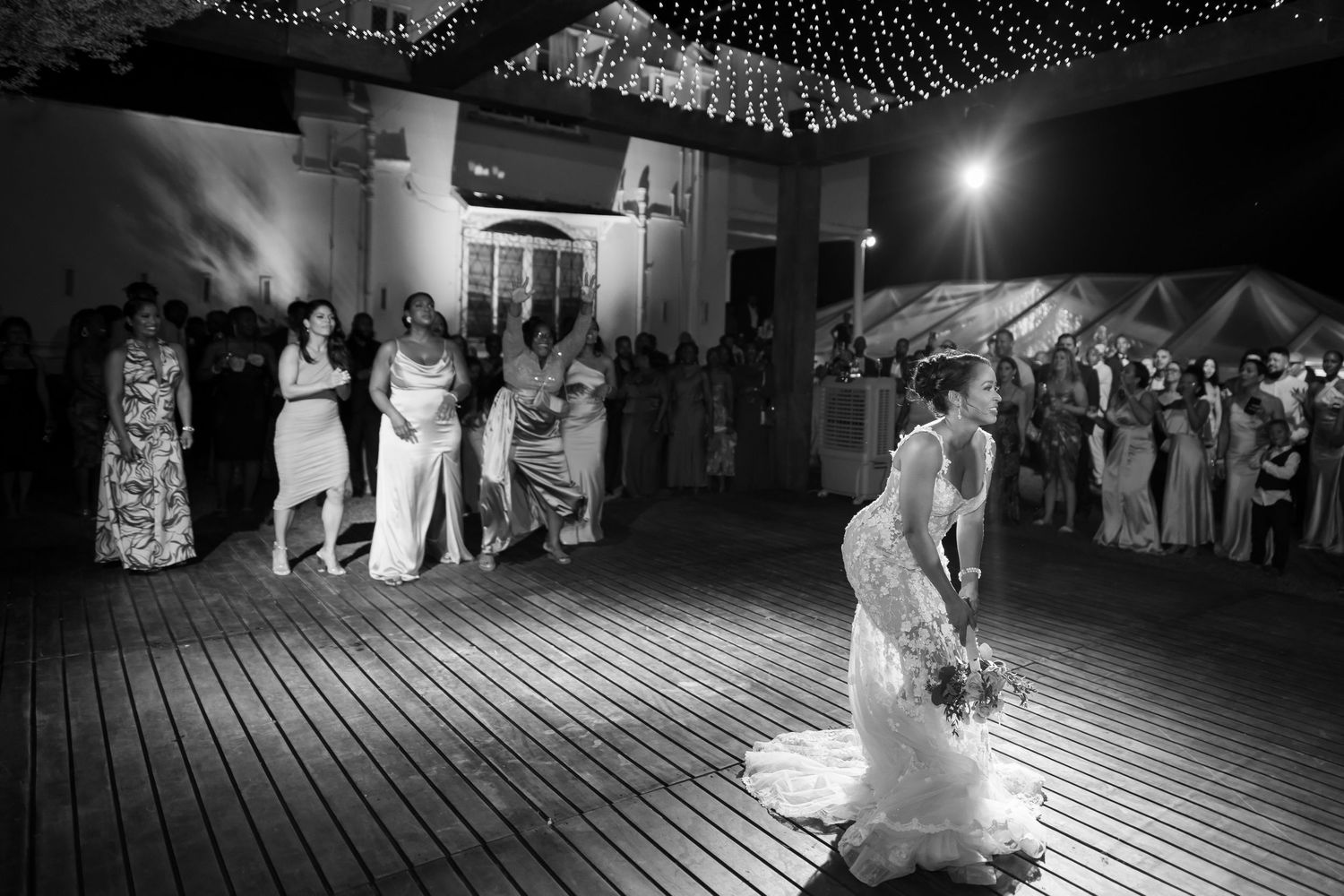 A black and white nighttime wedding reception scene on a wooden deck with guests gathered under twinkling lights.