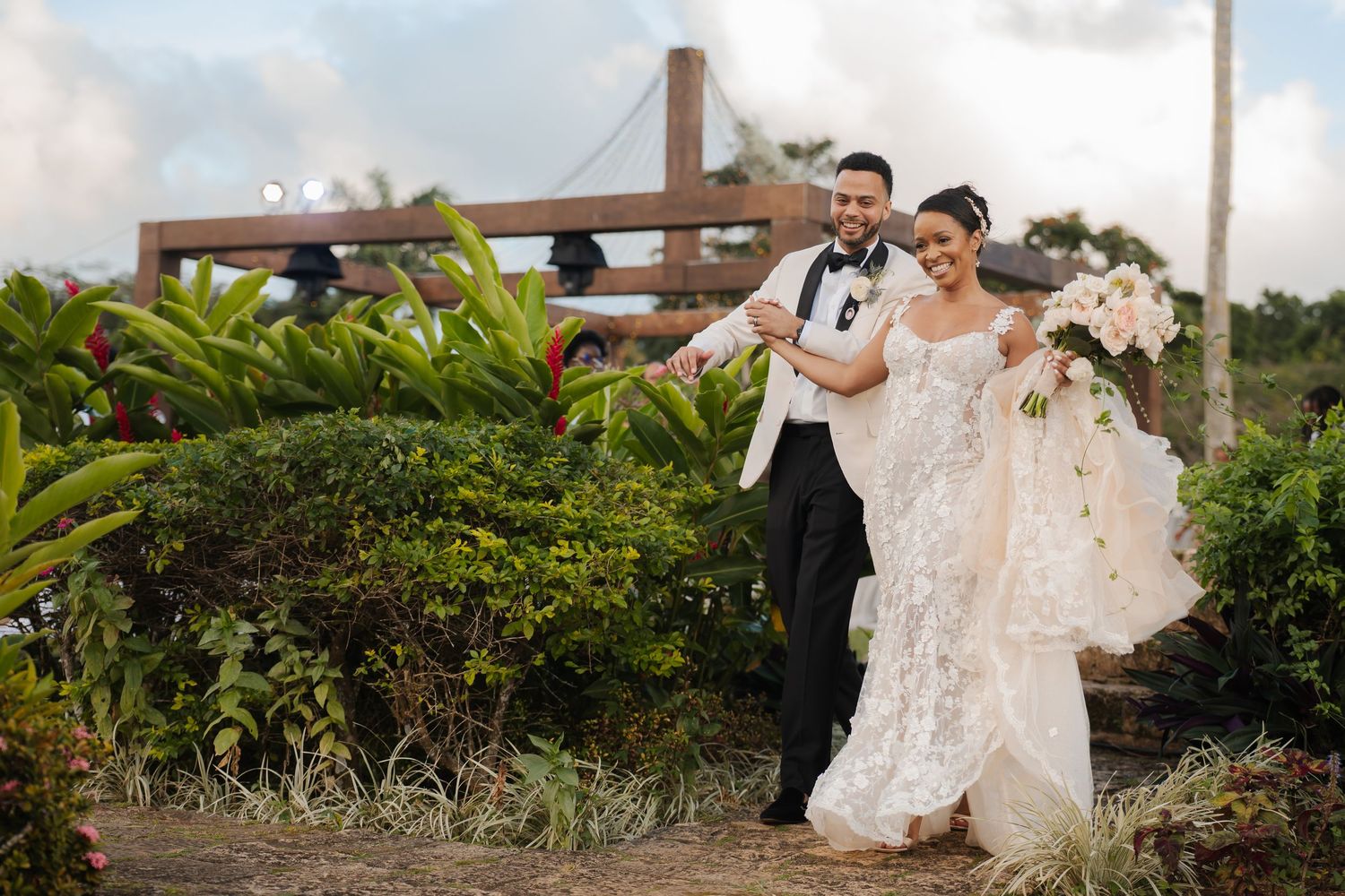 A couple shares a joyful moment on their wedding day in a tropical garden setting with a wooden cross backdrop.