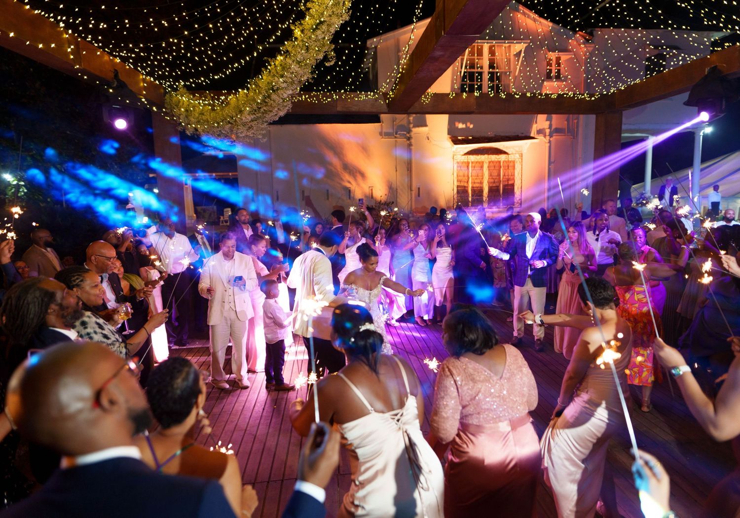 Wedding reception dance party with colorful blue and purple lighting under string lights on an outdoor deck.