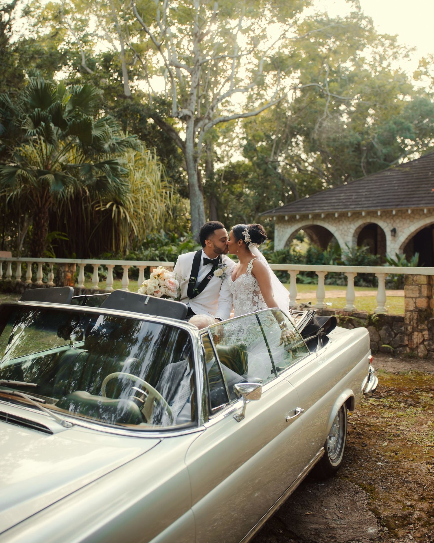 A classic cream-colored vintage convertible parked near arched architecture surrounded by lush tropical gardens.