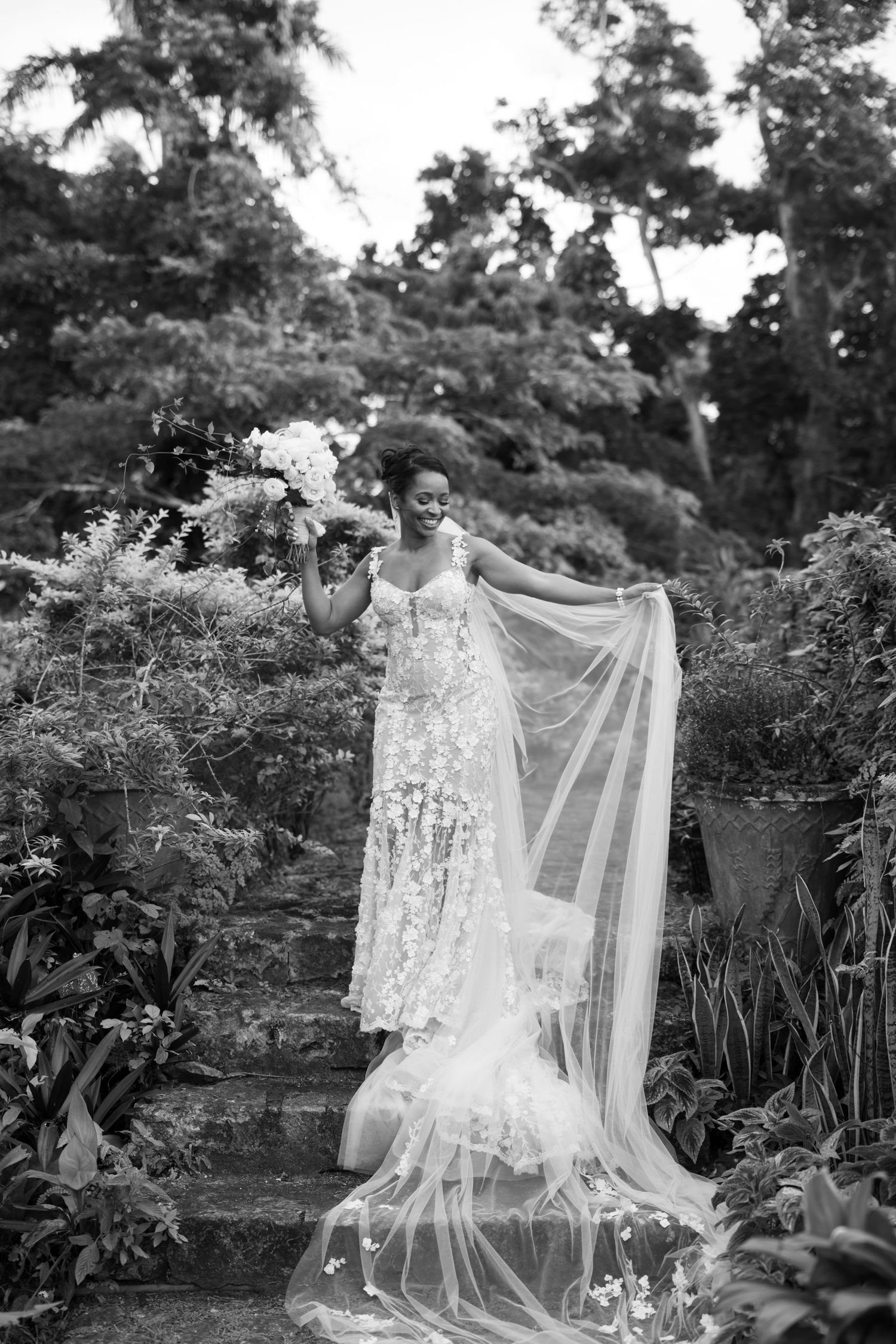 A bride in an elegant lace wedding gown with flowing veil stands gracefully on garden steps surrounded by lush foliage.