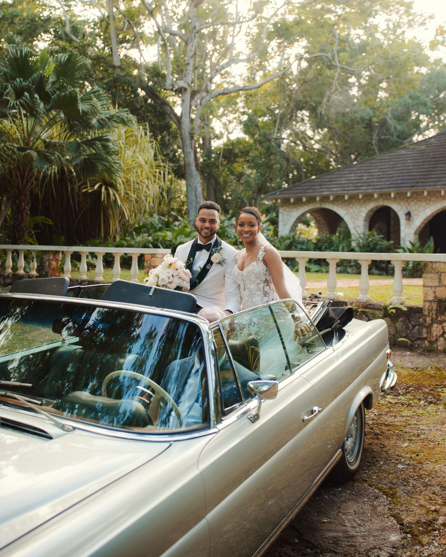 A classic vintage convertible parked by a Mediterranean-style building with tropical landscaping at sunset.