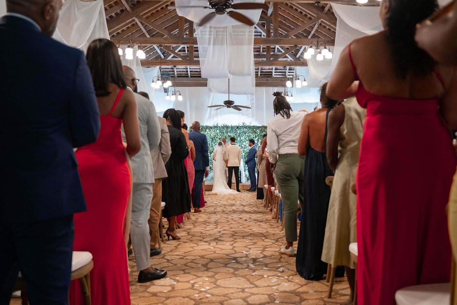 Wedding guests dance and socialize at a rustic reception venue with exposed wooden beams and string lights overhead.