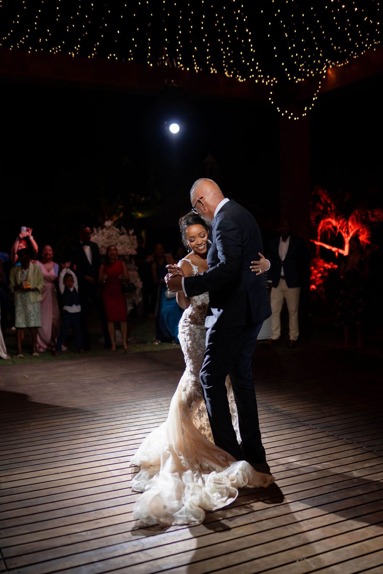 Couple performs romantic dip dance move under string lights on wooden deck at twilight wedding reception.