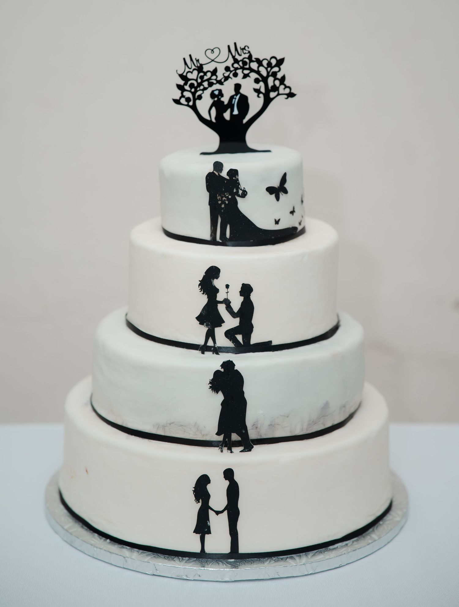 A four-tier white wedding cake decorated with black silhouettes showing a couple's love story from first meeting to marriage.