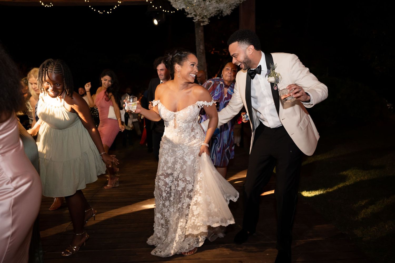 People dancing in a nighttime outdoor celebration with string lights and lush greenery creating a festive atmosphere.