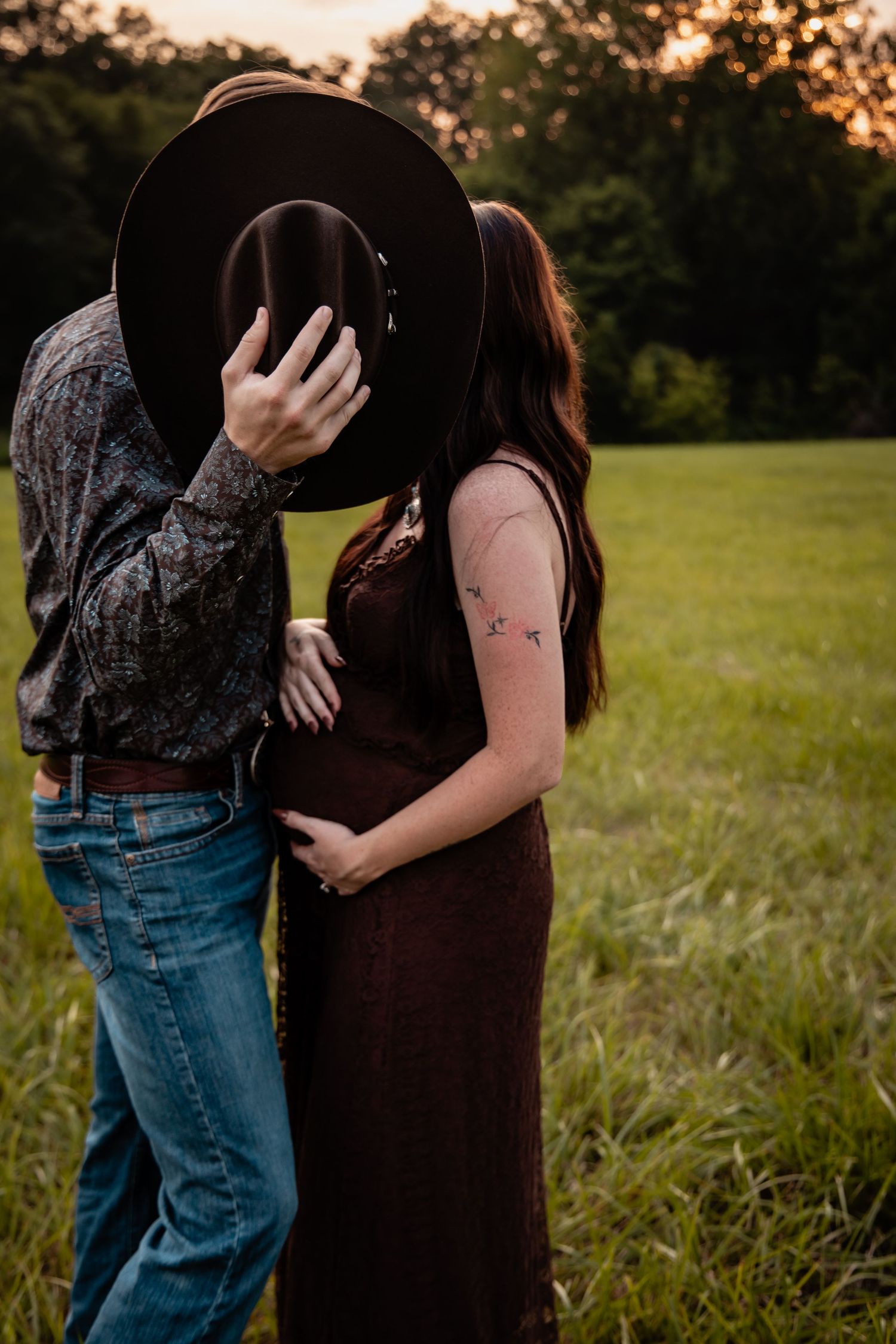 Couple sharing a kiss hidden behind a cowboy hat in a green field at sunset, woman in burgundy dress showing baby bump.