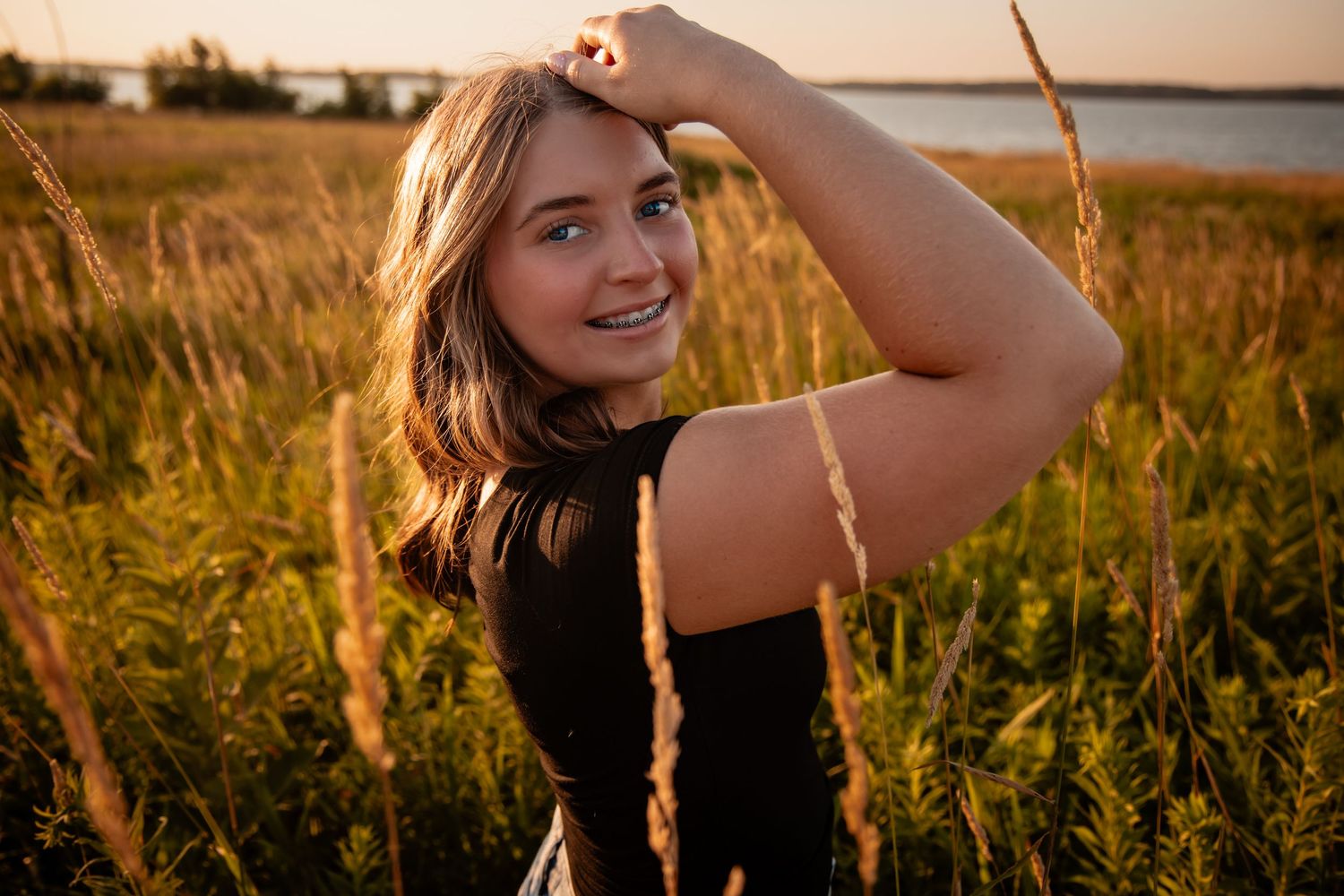 Young woman smiling in golden hour sunlight among tall grass near a lake, wearing a black top.
