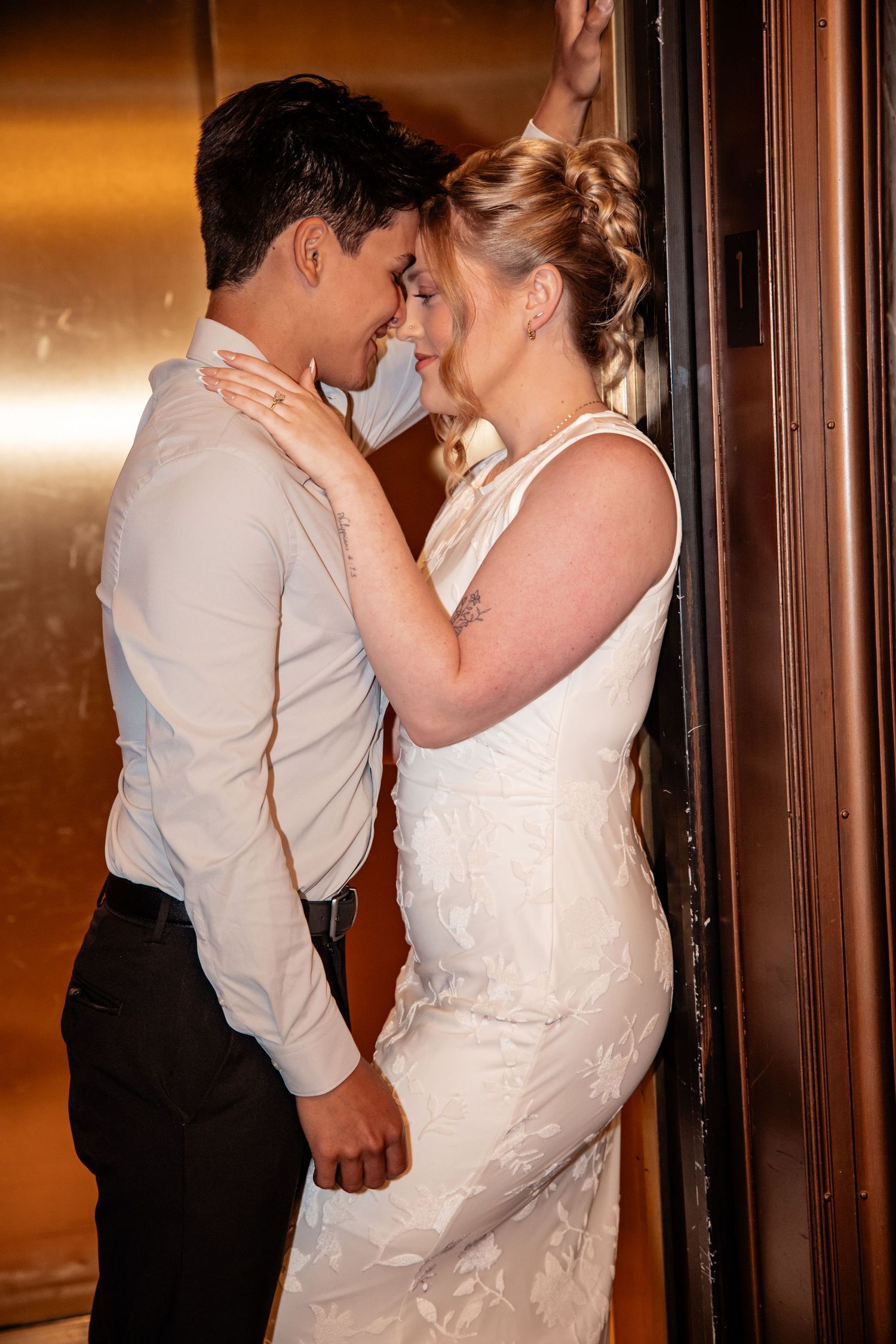 A bride and groom share an intimate moment in a golden elevator, dressed in wedding attire, foreheads nearly touching.