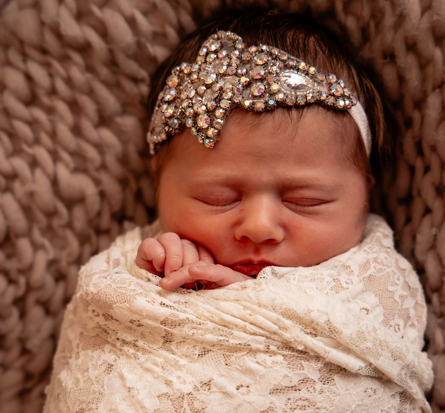 Sleeping newborn baby swaddled in lace wrap wearing a rhinestone headband on a knit blanket.
