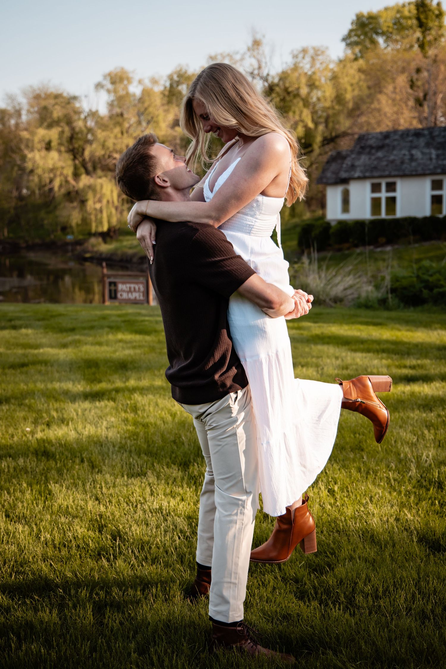 A man lifts a woman in a white dress joyfully in a green field near a chapel at sunset.