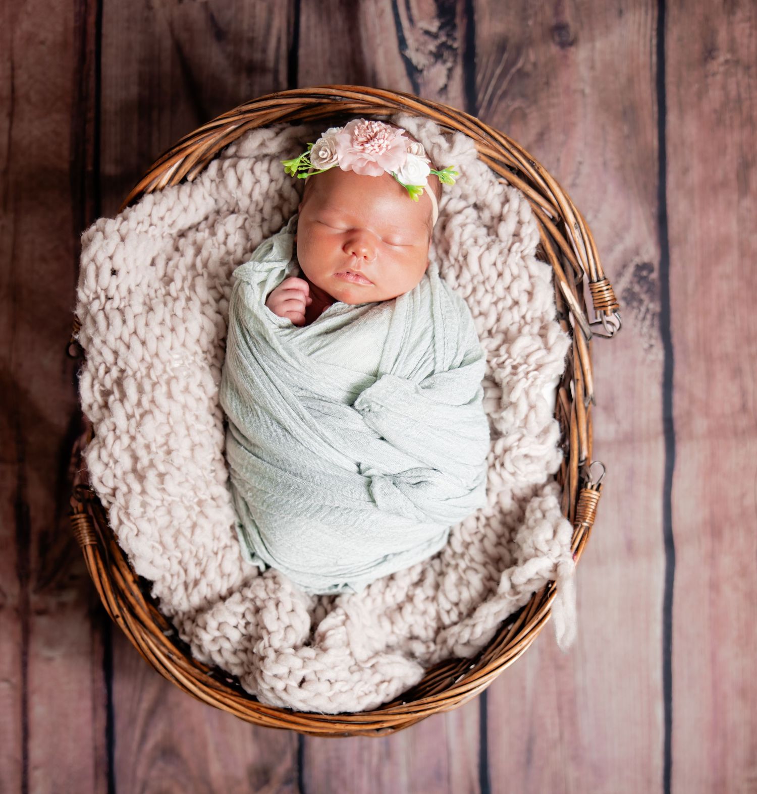 Newborn baby girl swaddled in mint wrap with floral headband sleeping peacefully in a wicker basket.