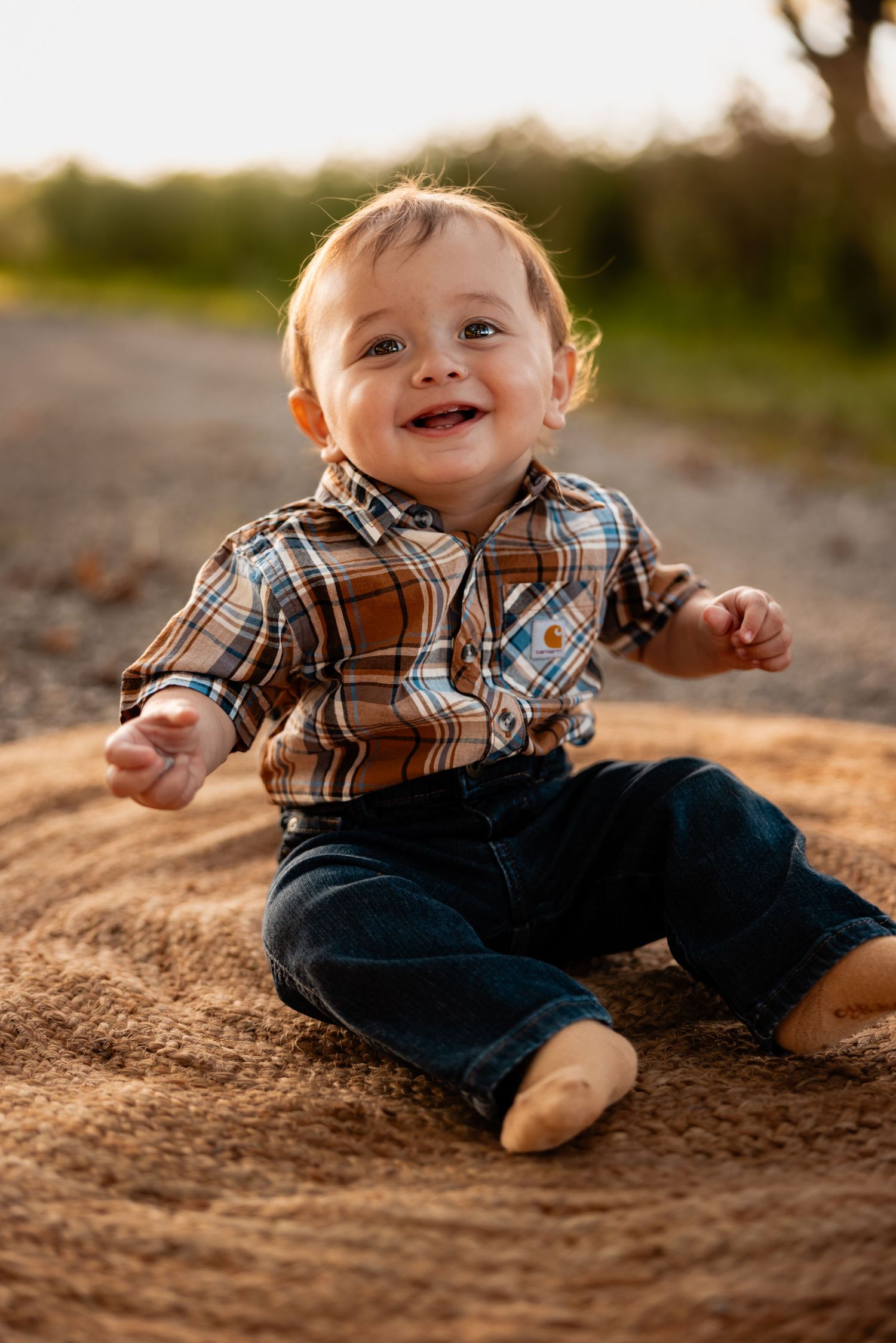 Smiling baby boy in plaid shirt and jeans sitting outdoors on hay bale in golden sunset light.