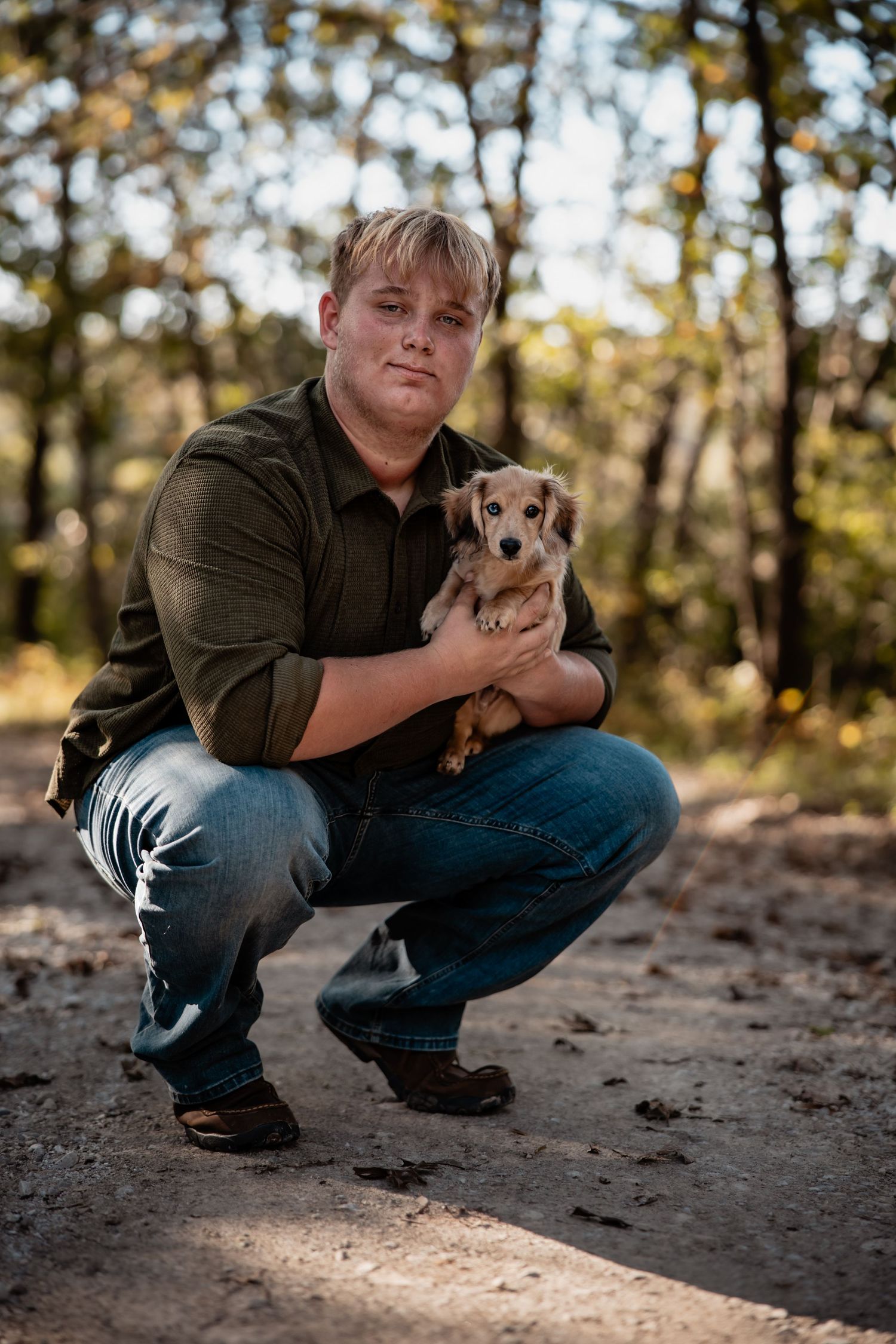 Man crouching outdoors holding a small dachshund puppy in a wooded area wearing a green shirt and jeans.