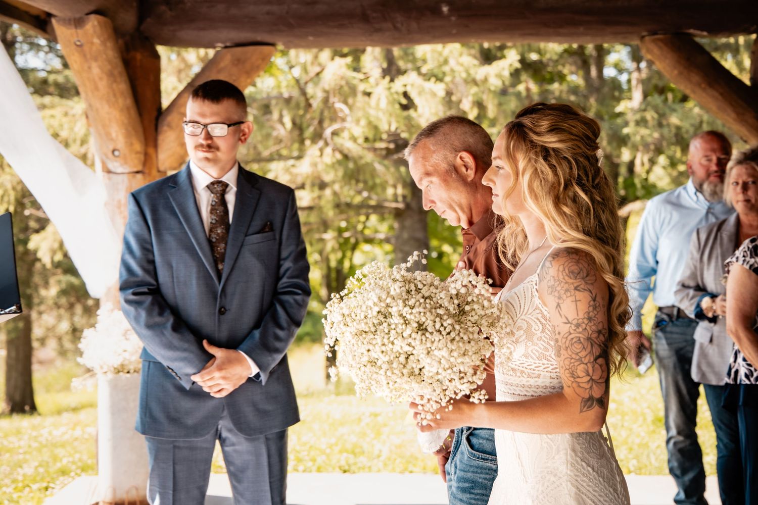 Outdoor wedding ceremony with bride holding baby's breath bouquet, groom kissing her forehead, officiant watching nearby.