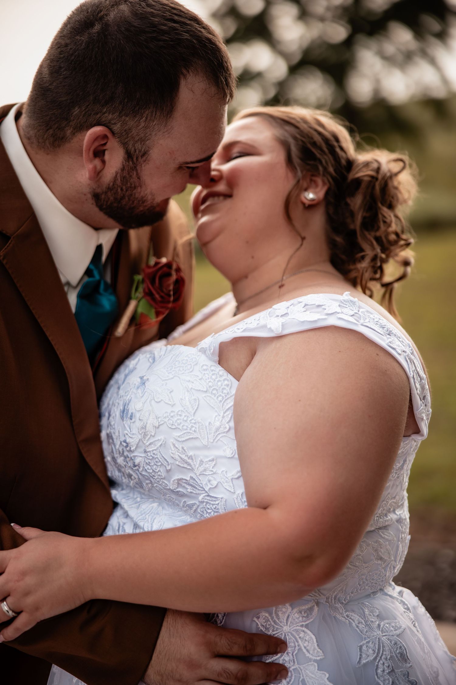 A bride and groom share a tender nose-to-nose moment, she smiling in white lace, he in a brown suit with teal tie.