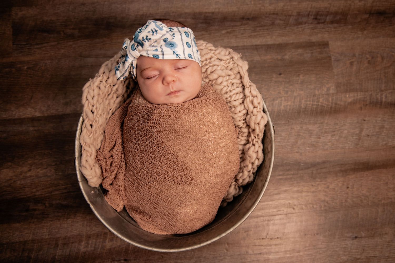 Newborn baby swaddled in brown wrap, sleeping in a bowl with knit blanket and floral hat.