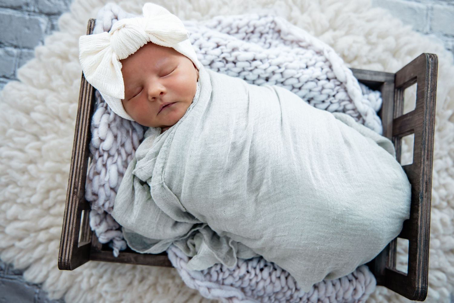 Newborn baby swaddled in light blue wrap with white bow headband sleeping in small wooden crib on knit blanket.