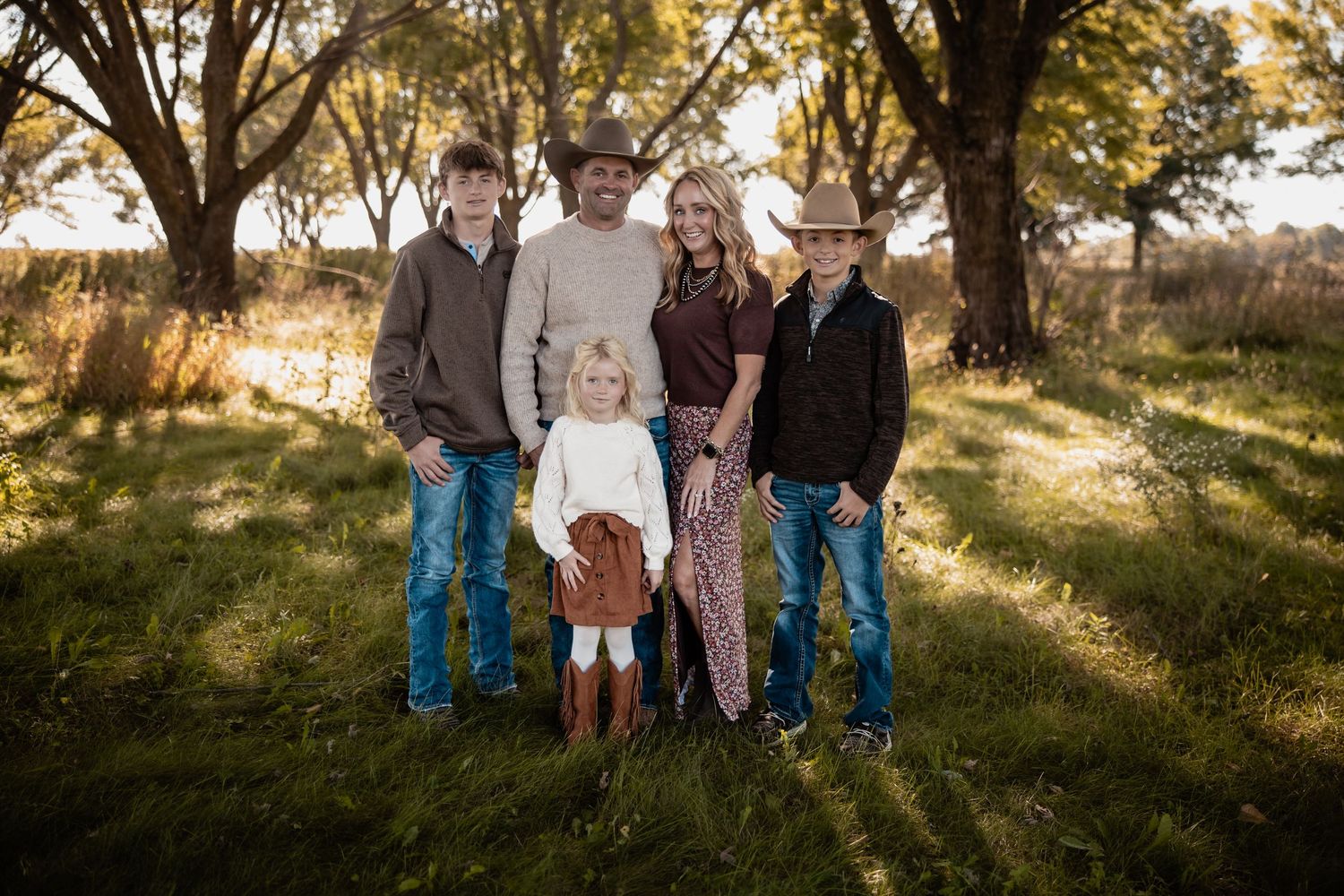 Family of five posing outdoors in autumn, wearing western attire, surrounded by golden trees and green grass.