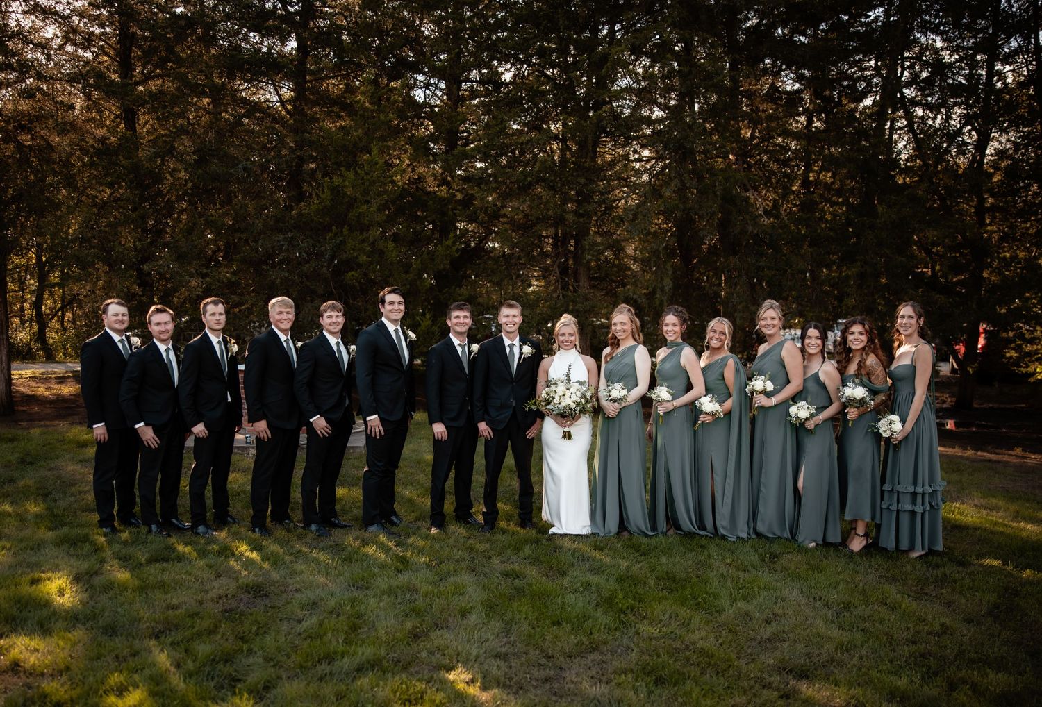 Wedding party in black suits and sage green dresses posing outdoors near trees.