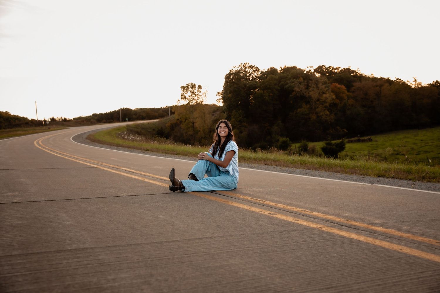 Young woman in blue dress sitting on a winding rural road at sunset with trees in background.