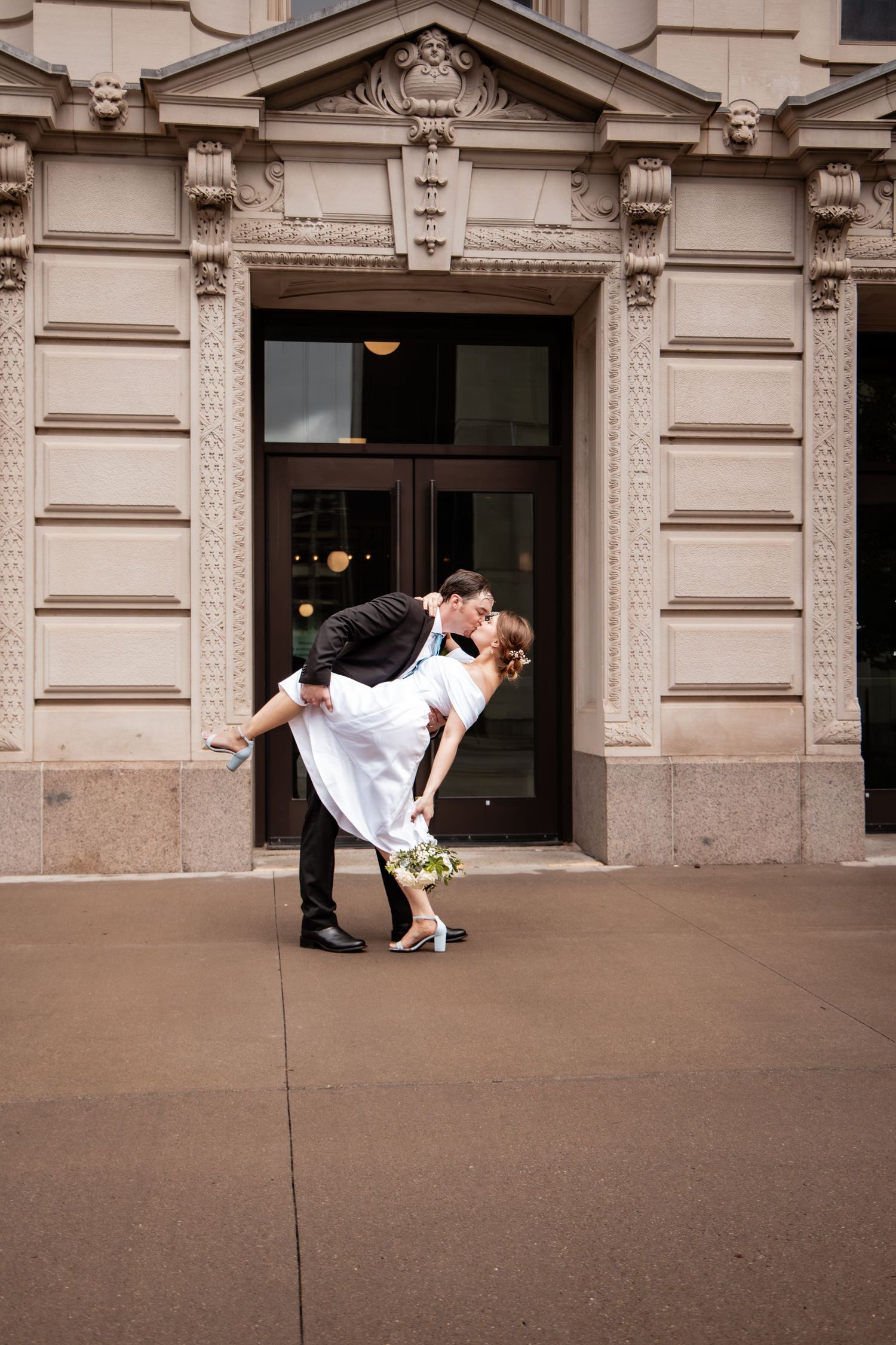 Groom dips bride for a romantic kiss outside an ornate beige stone building on their wedding day.