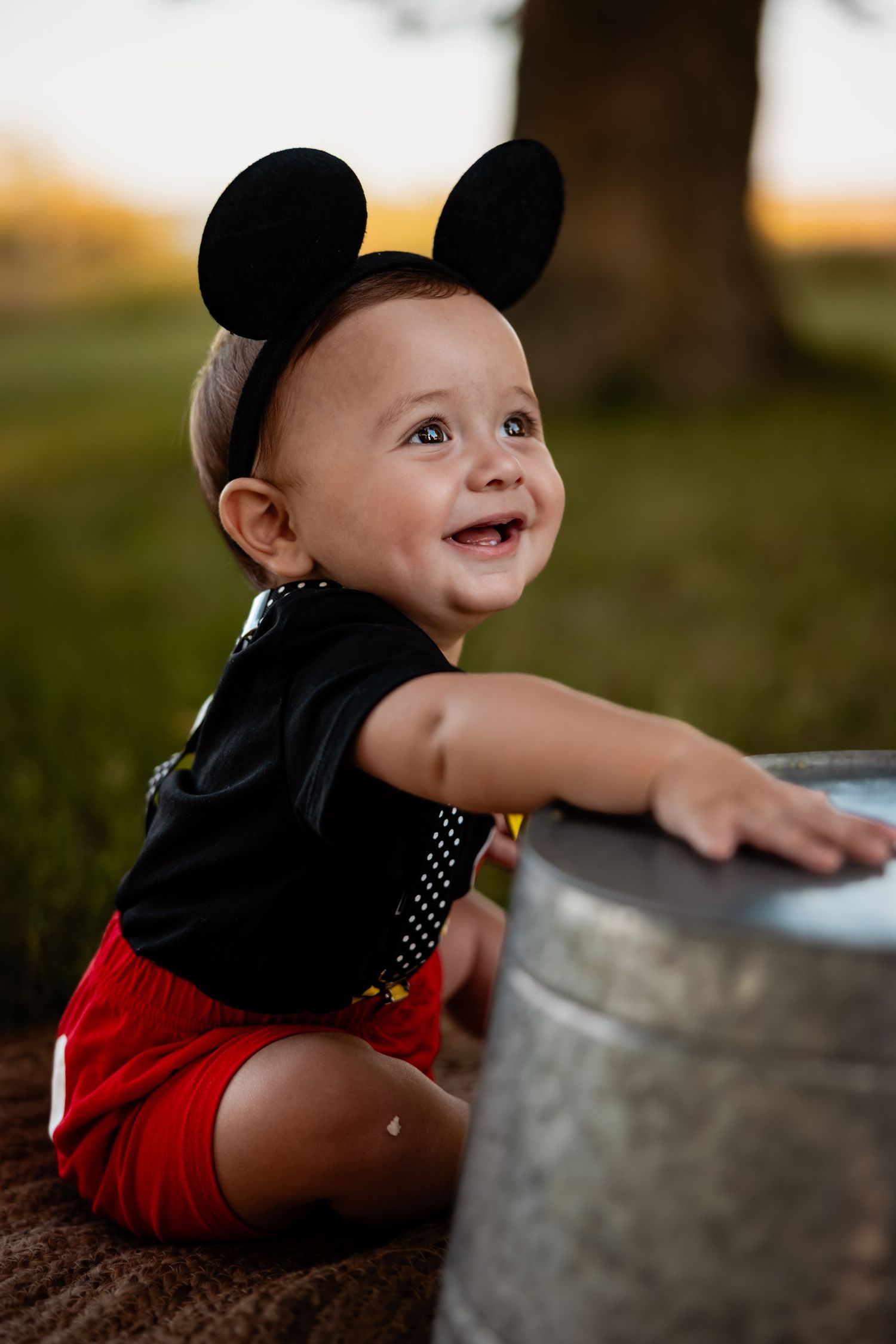 Smiling baby in Mickey Mouse ears headband and red shorts kneeling outdoors near a metal bucket.