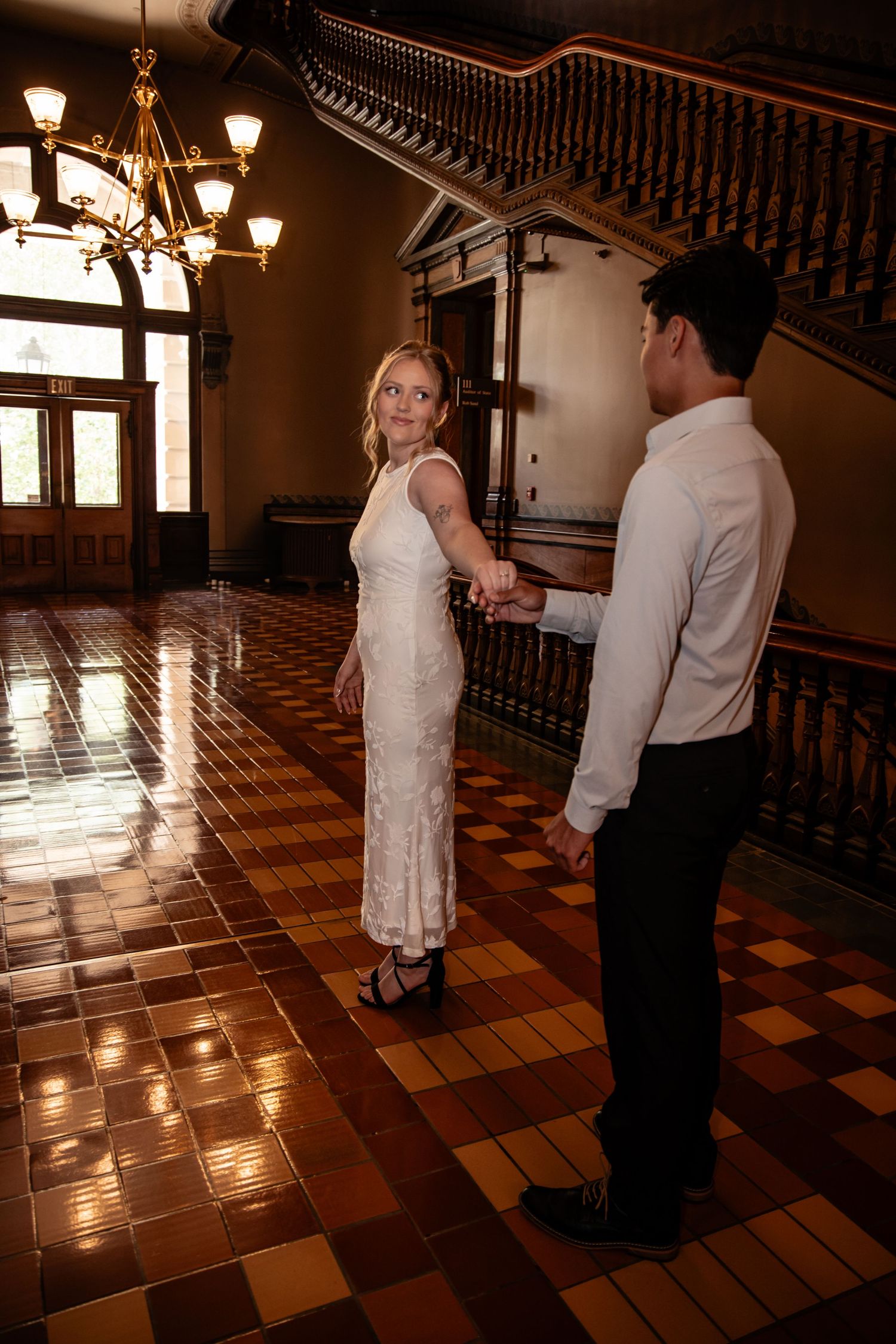 Couple holding hands in elegant historic building with chandelier and checkered tile floor, woman in white dress looking back.