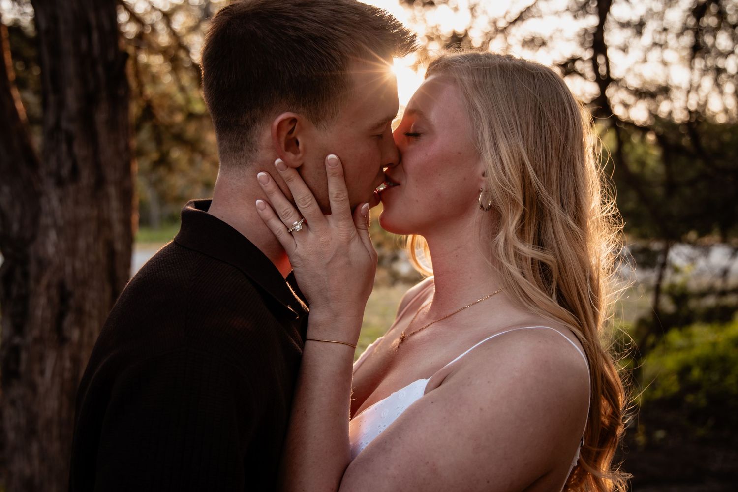 An engaged couple shares a golden sunset kiss with warm backlight filtering through trees behind them.