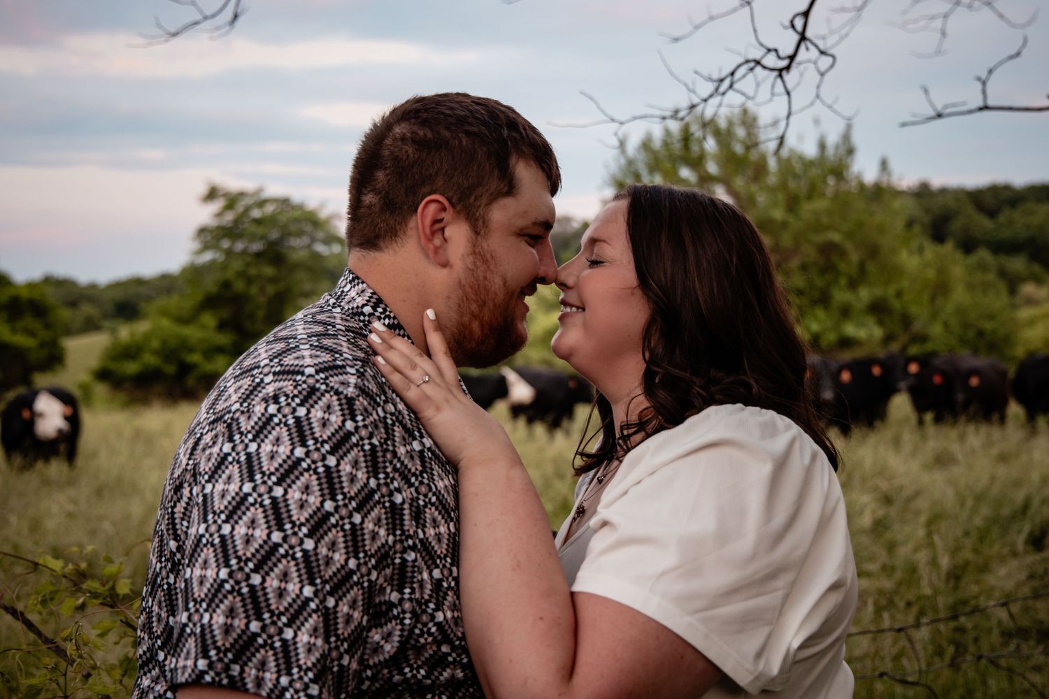 Couple about to kiss in a green field with cattle grazing in background.