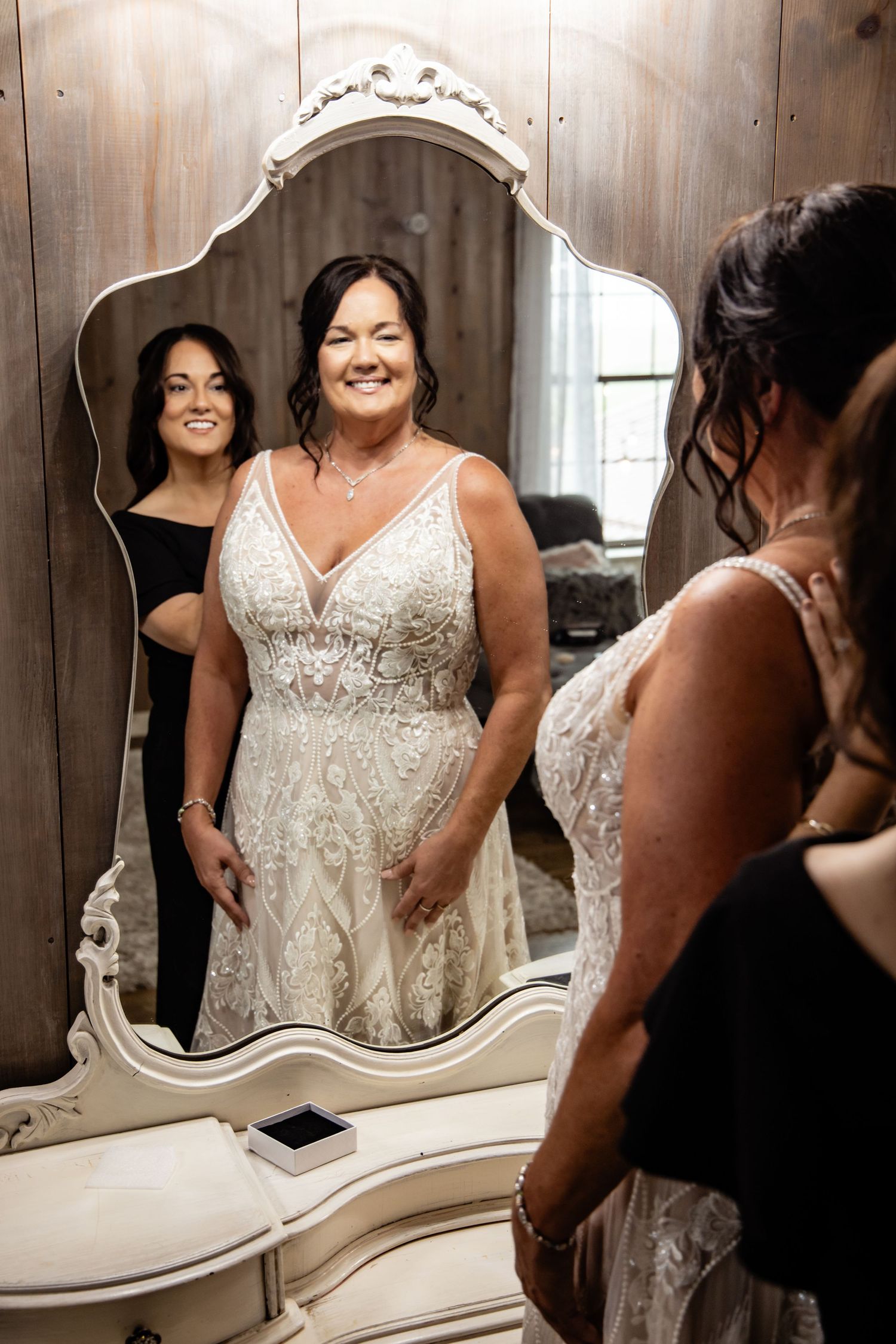 Bride in elegant beaded wedding gown smiling at her reflection in an ornate white mirror with bridesmaid nearby.
