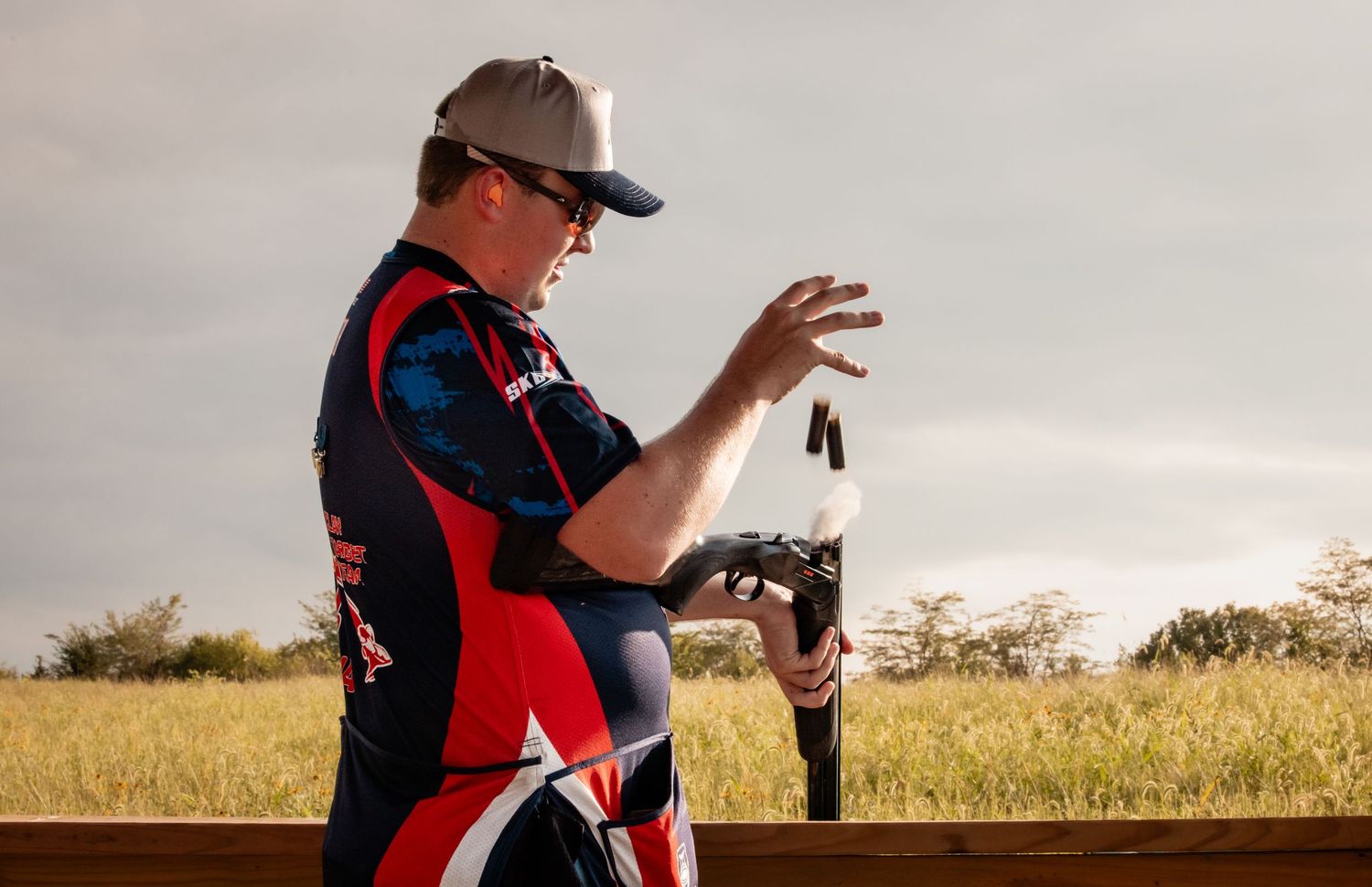 Man in USA shooting sports jersey loading a shotgun at an outdoor range with golden fields in background.