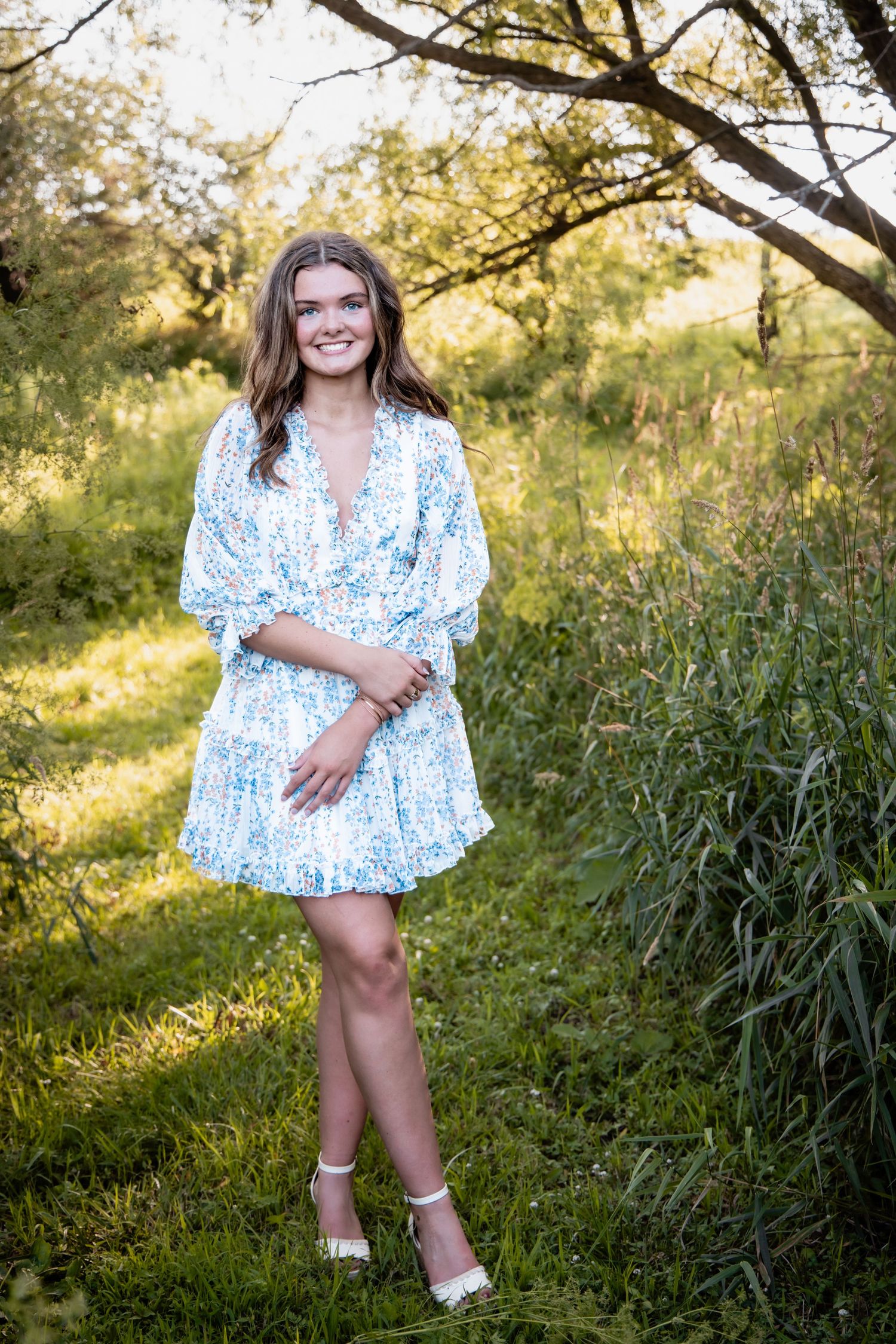 Young woman in white floral dress smiling outdoors in lush green nature setting during golden hour.
