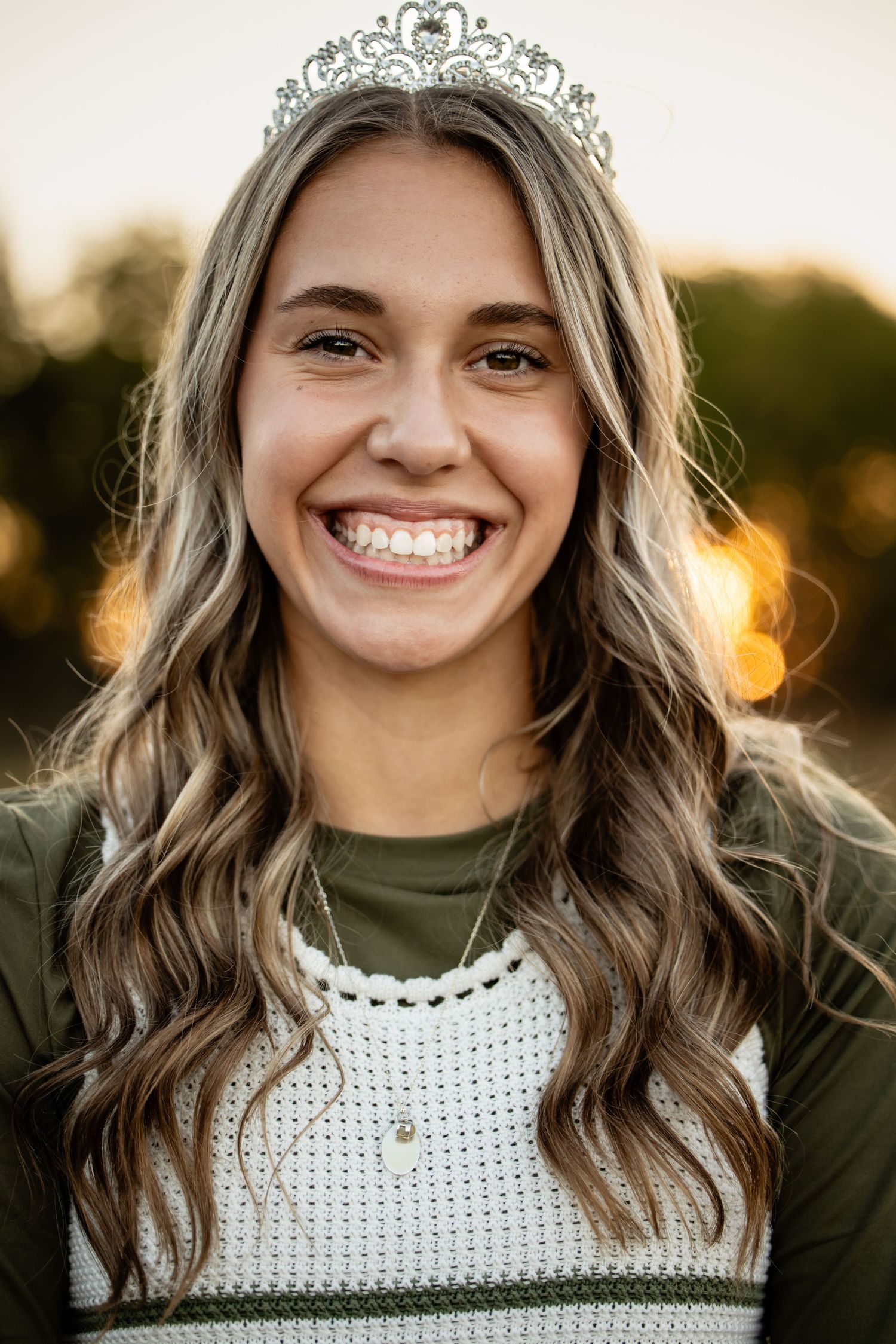 Smiling young woman wearing a sparkling tiara and green outfit with golden sunset bokeh background.