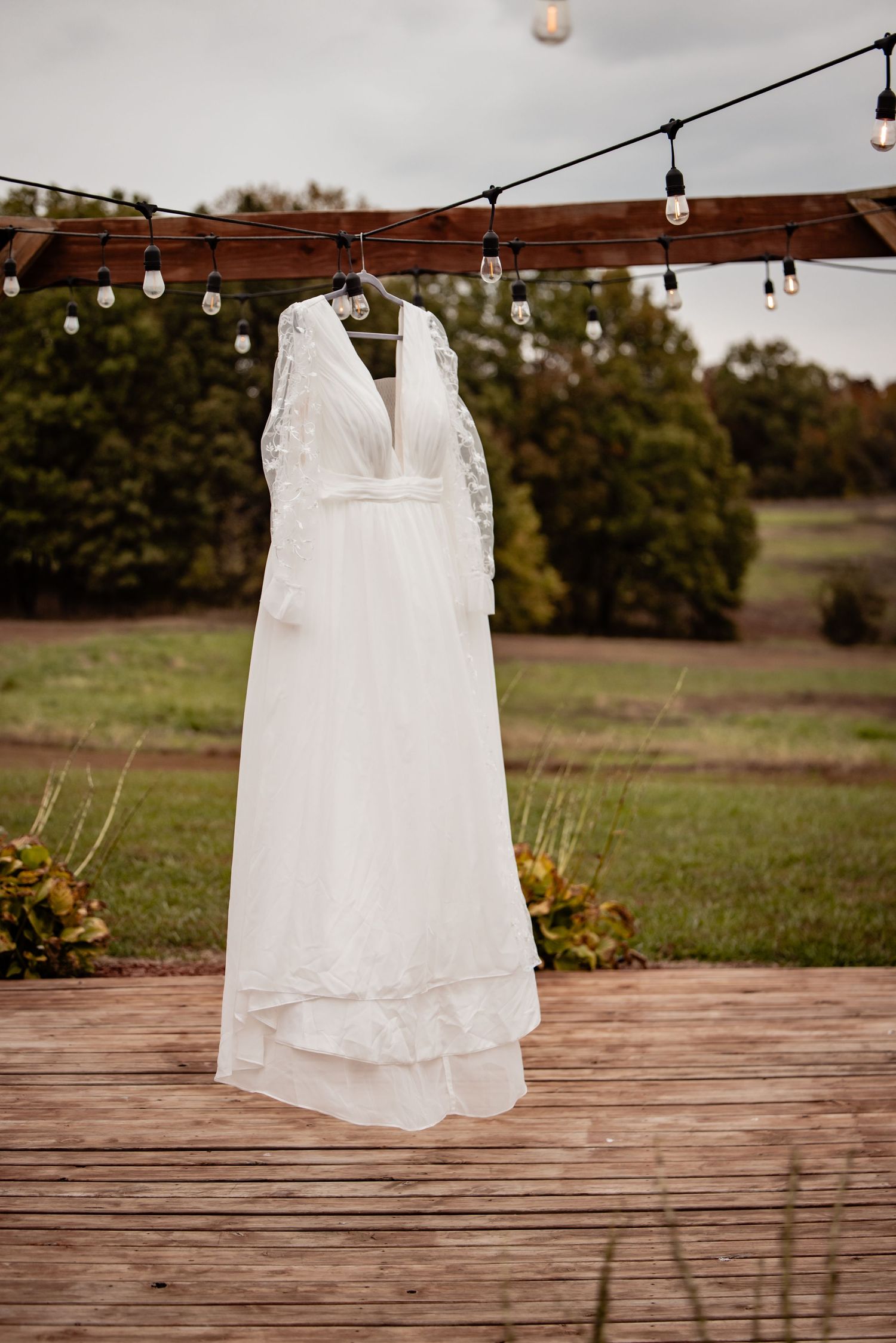 Elegant white lace wedding dress hanging on a wooden pergola with string lights outdoors.