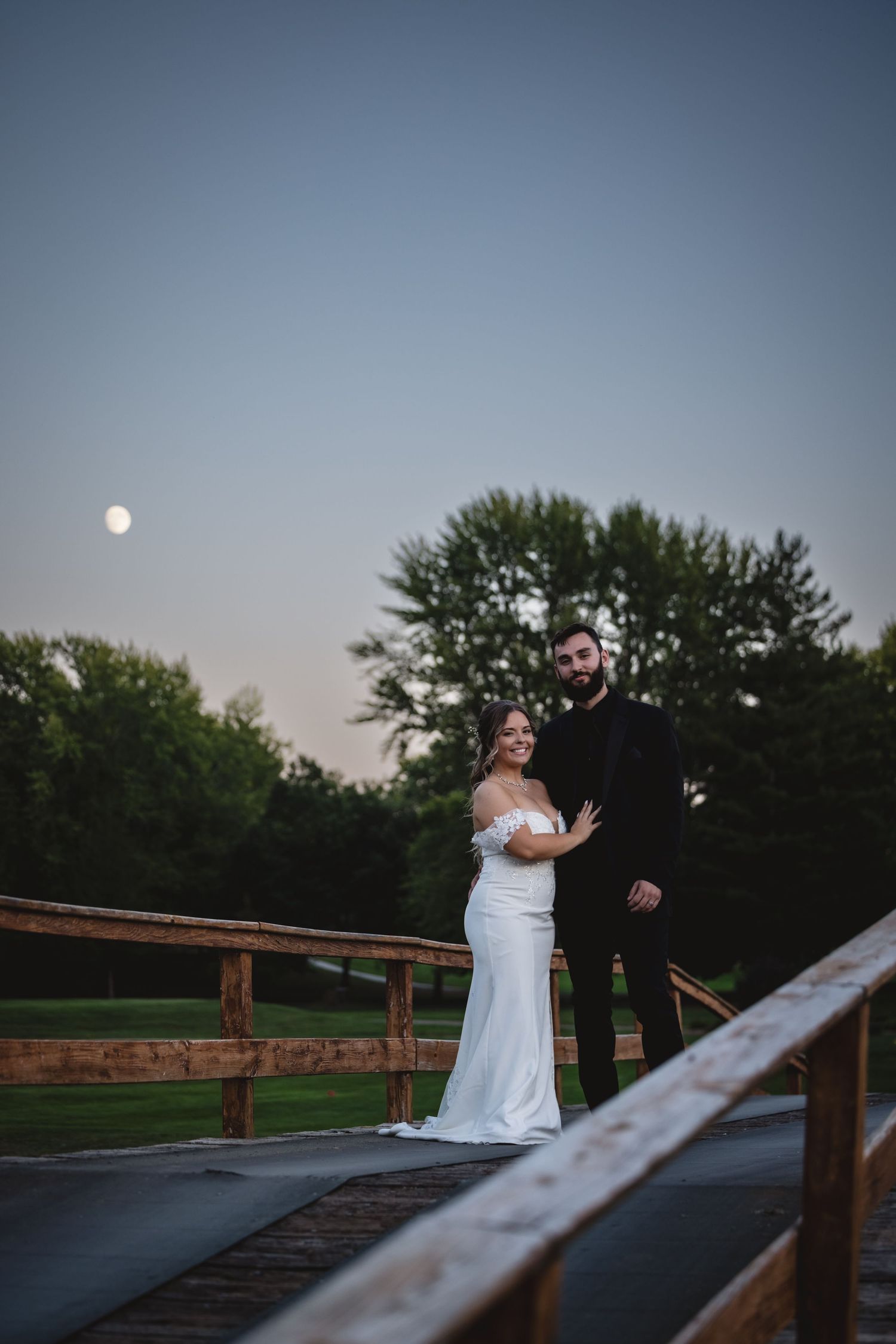 Bride and groom pose on a rustic wooden bridge under a full moon at dusk.
