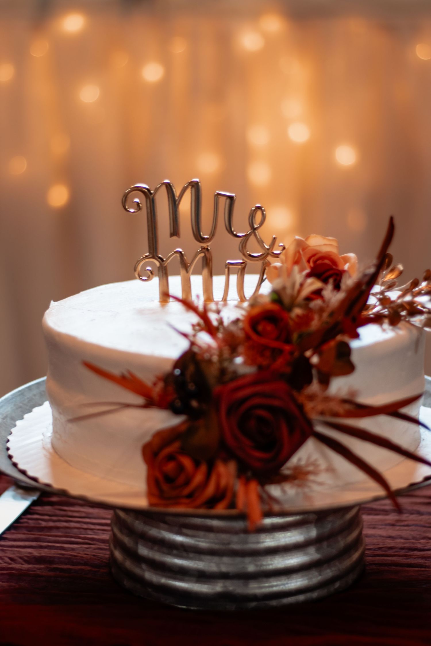 White wedding cake with Mr & Mrs topper and autumn floral arrangement on rustic stand with bokeh lights.