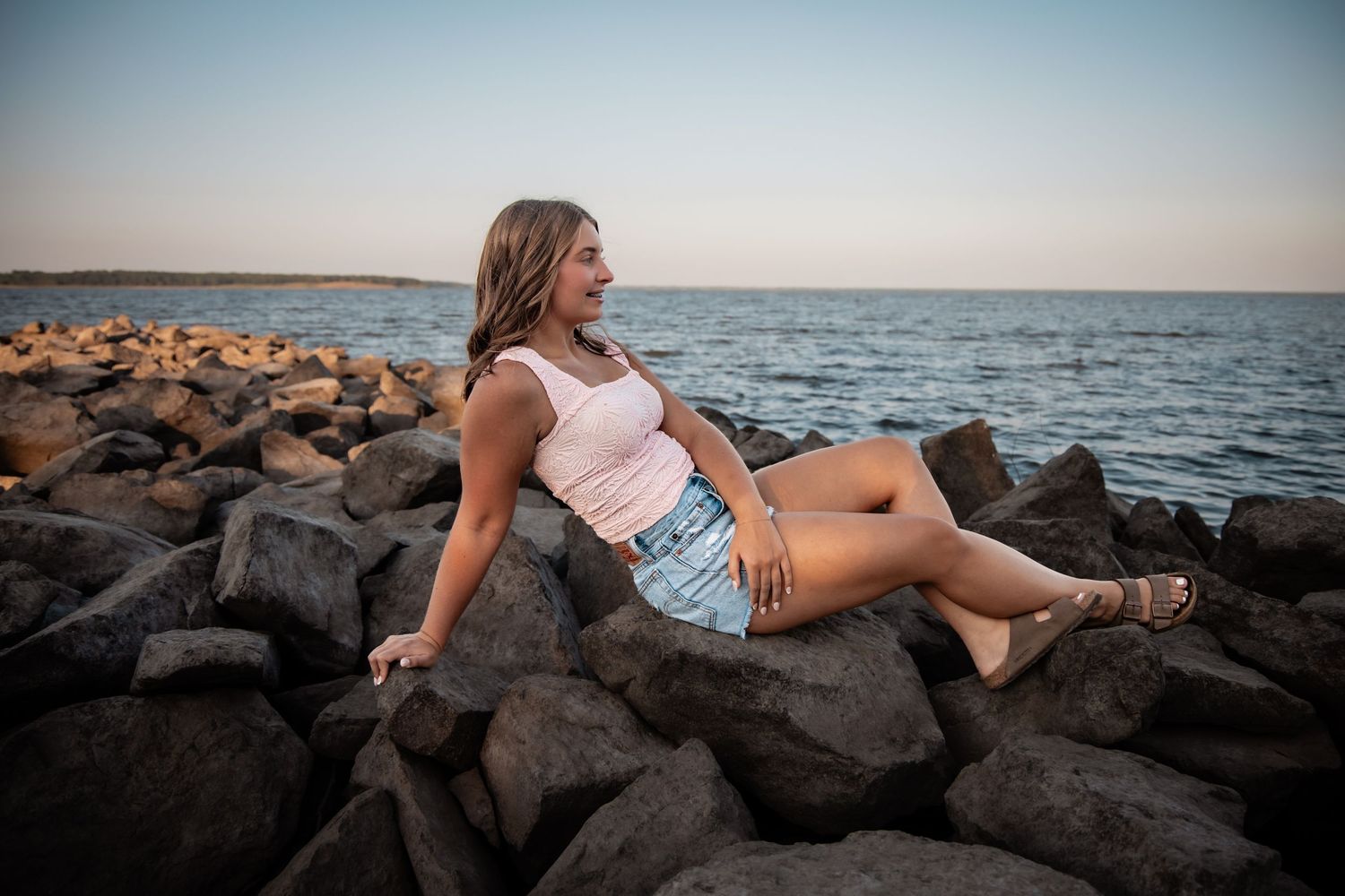 Young woman in pink top and denim shorts sitting on rocky shoreline at sunset with calm ocean in background.