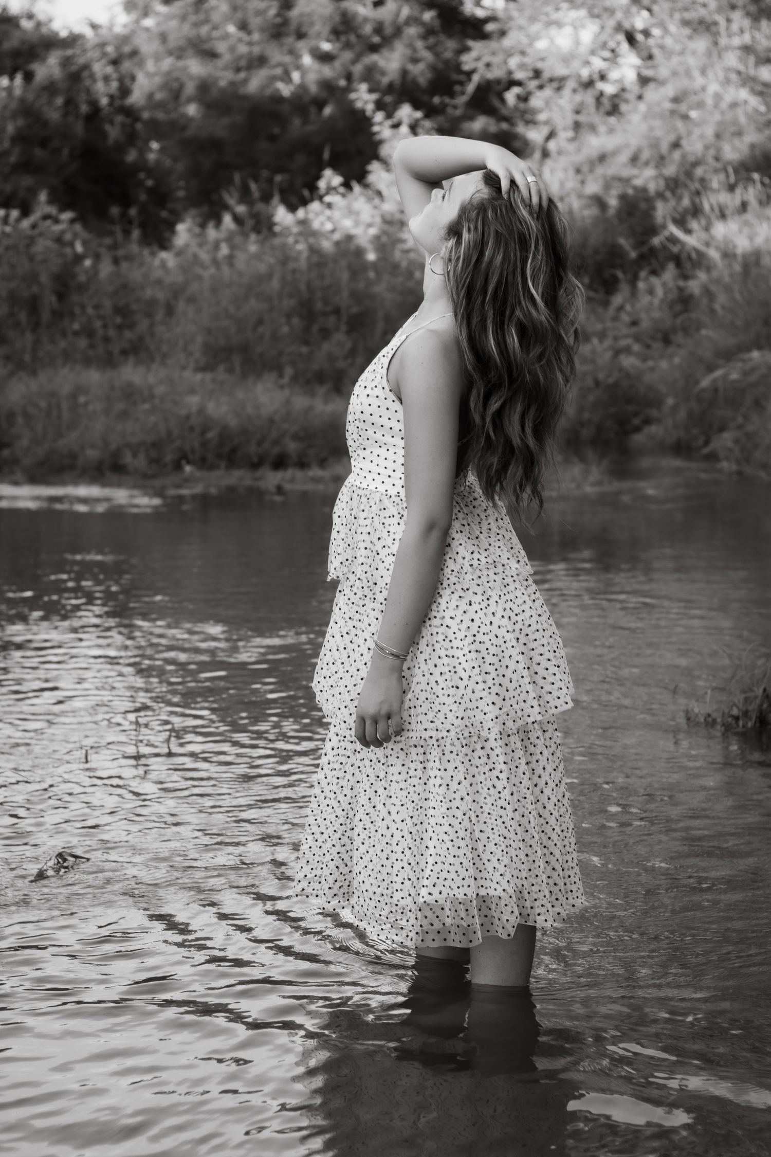 Black and white photo of a young woman in a polka dot dress standing in a shallow river surrounded by lush greenery.